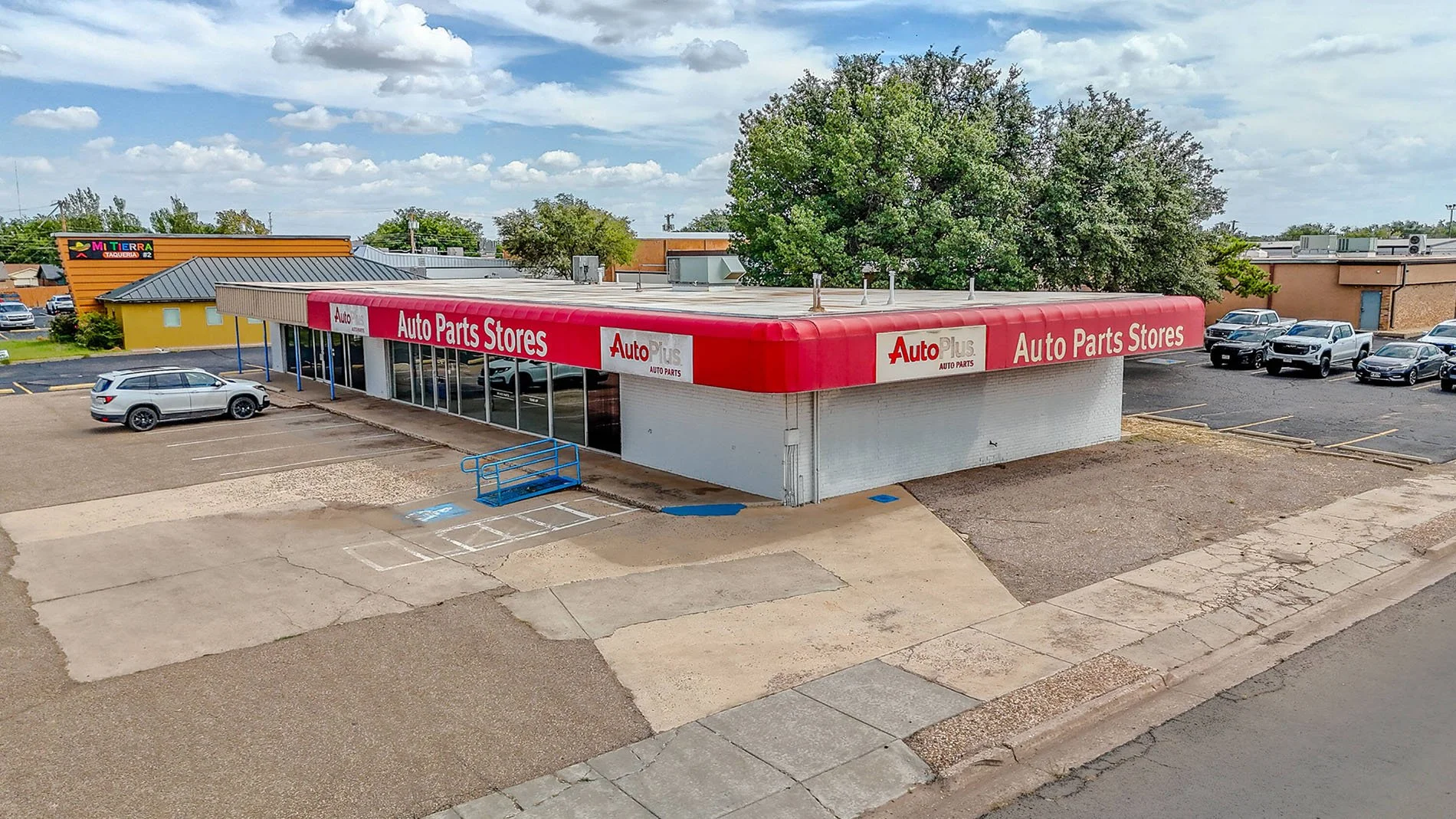An auto parts store with a red awning sign reading 'Auto Parts Stores' and 'Auto Plus', located in a small commercial area with parking spaces in front, a tree in the background, and partly cloudy sky.
