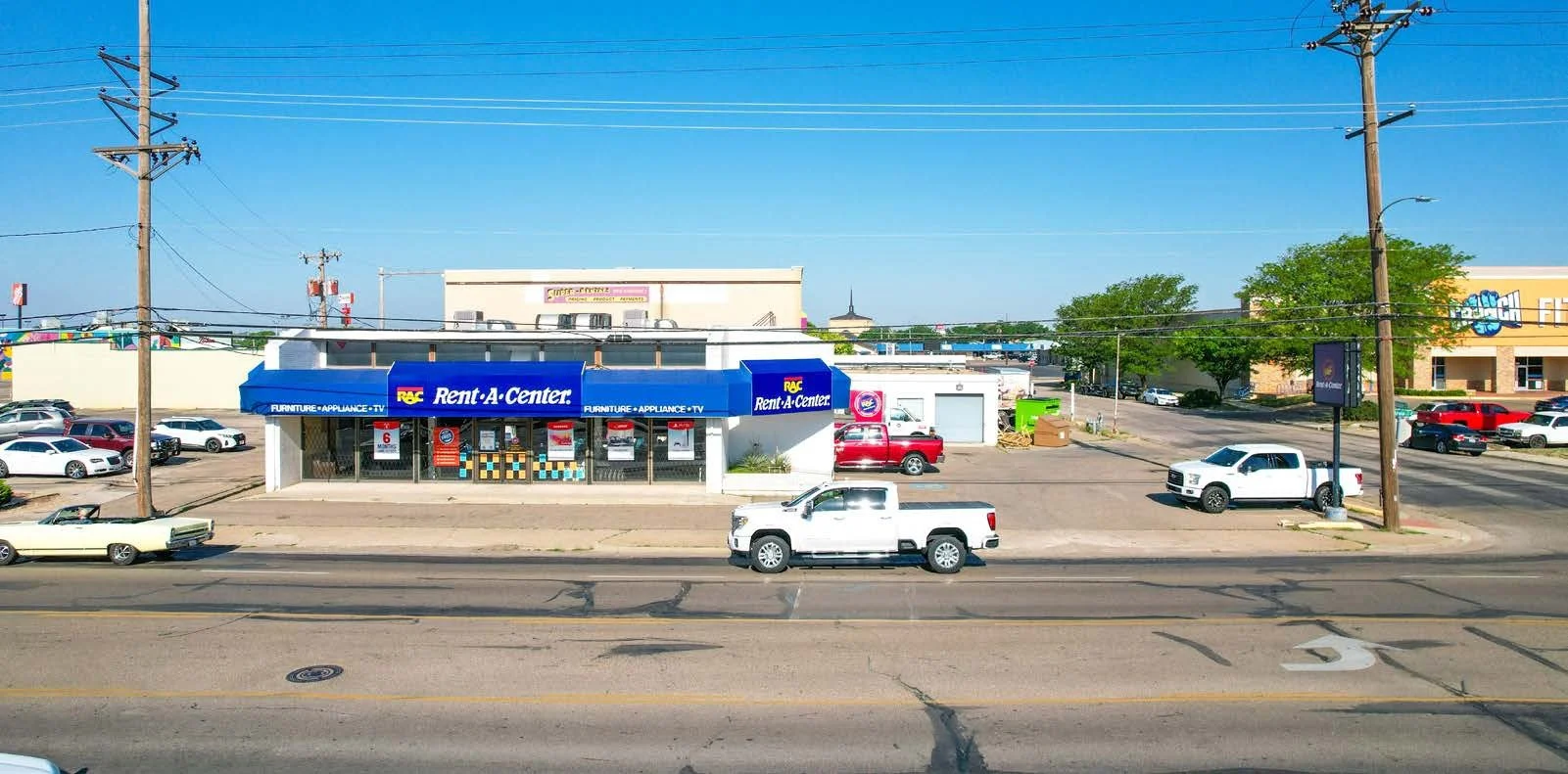 A storefront with a blue awning displaying 'Rent-A-Center' and a parking lot with several cars, utility poles, and a clear blue sky.