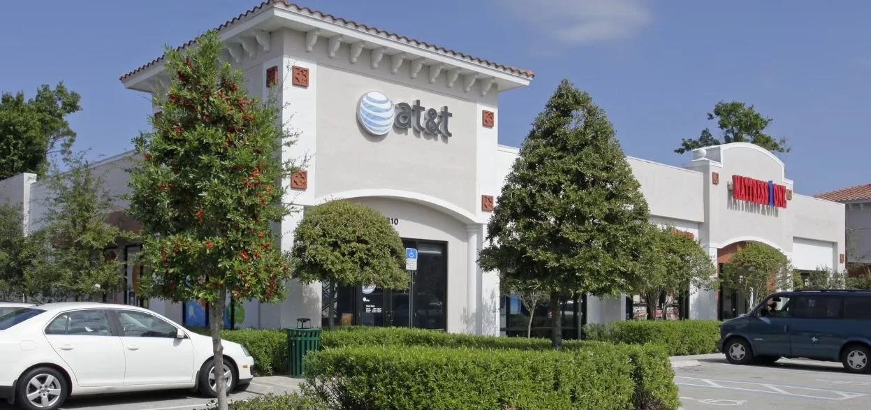 A shopping plaza with an AT&amp;T store and a Mattress One store, parking lot with cars, and trees in front.