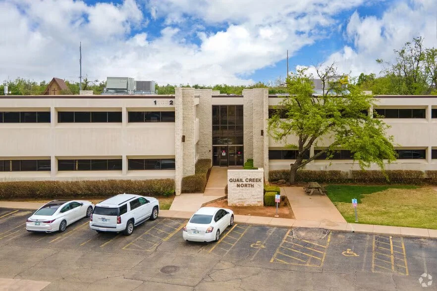 Multi-story office building labeled 'Quail Creek North' with a parking lot and several parked cars, trees, and a partly cloudy sky.