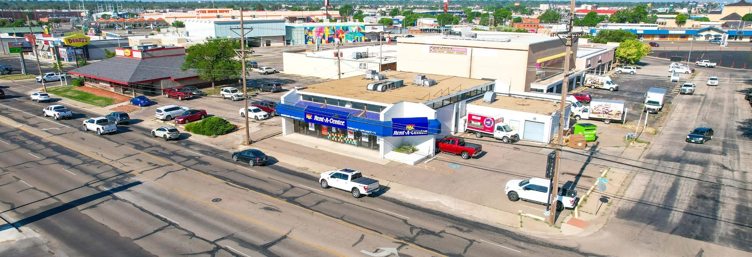 Aerial view of a shopping area with multiple stores, parking lot, and cars on the street. Notable storefront is a Rent-A-Center in the foreground. Shops include fries-frites, Denny's, and others with colorful signage. Parking lots mostly empty and gr