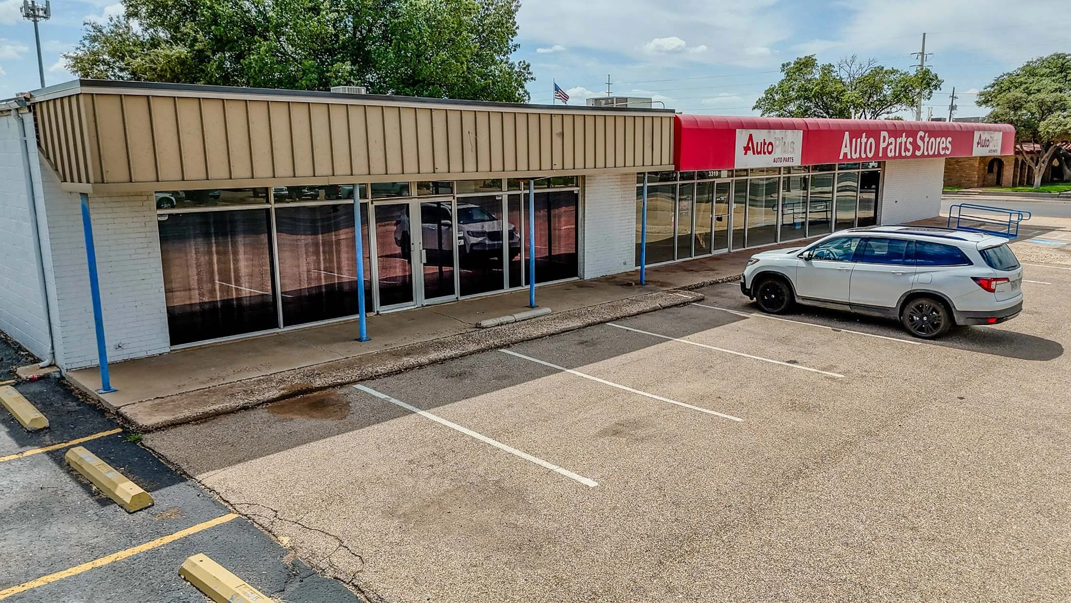 An auto parts store with a red awning labeled 'Auto Parts Stores', a beige exterior, and empty parking spaces in front. A white SUV is parked next to the store with a blue cart on top.