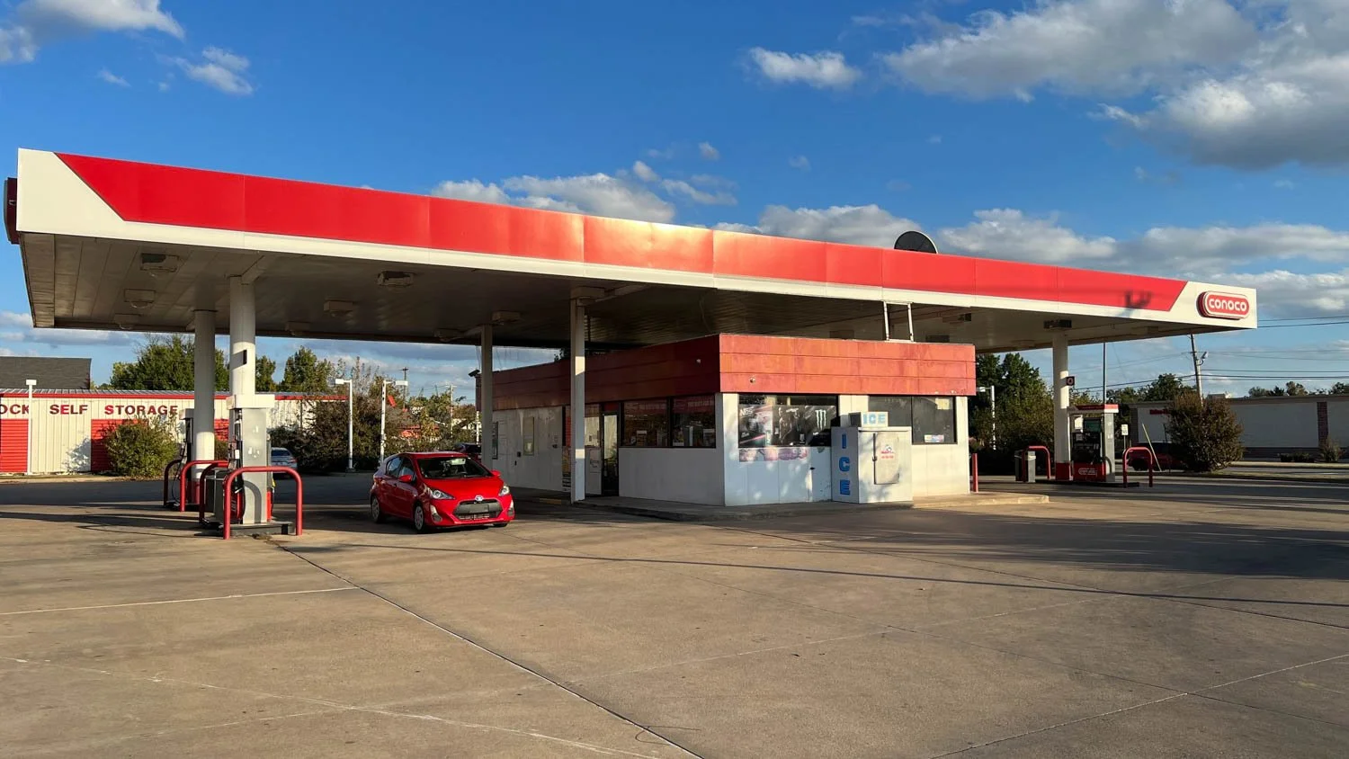 Conoco gas station with red and white canopy, red car parked at pump, ice vending machine, and self-storage building in the background under partly cloudy sky.