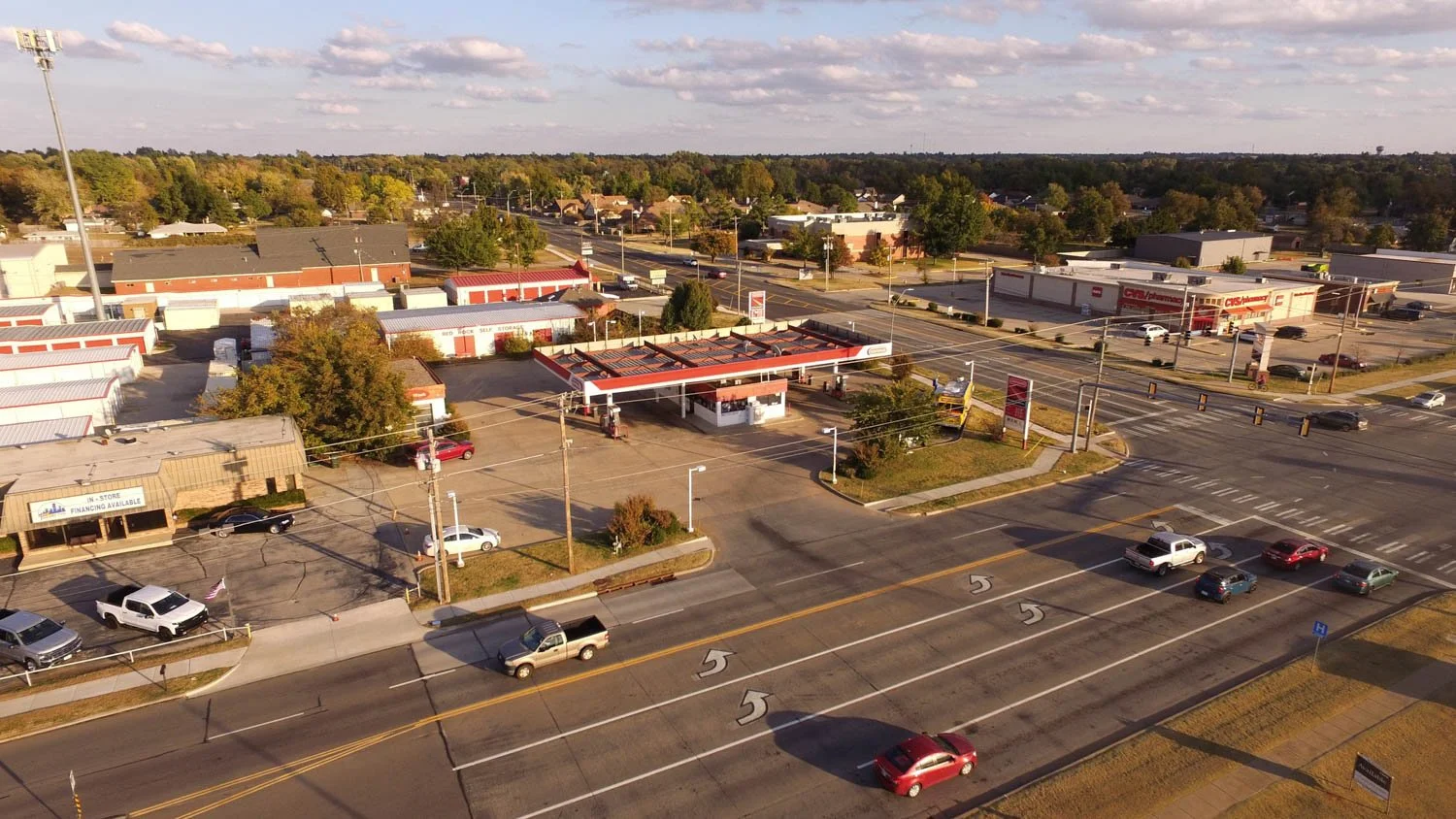 Aerial view of a small commercial area with a drive-thru, restaurants, retail stores, scattered cars, and traffic lights on a clear day.