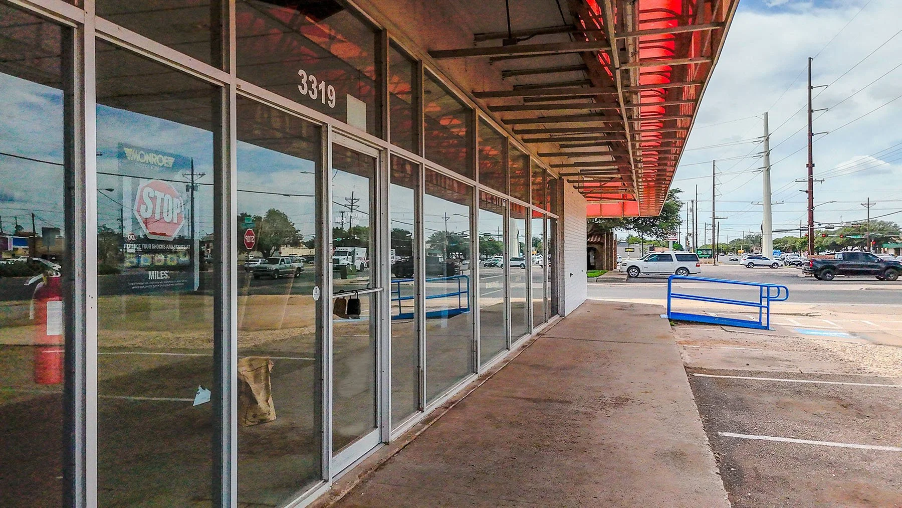 Empty storefront with glass windows and a glass door, located at 3319, with blue bike racks outside on a sidewalk. The parking lot has several cars and power lines are visible in the background.
