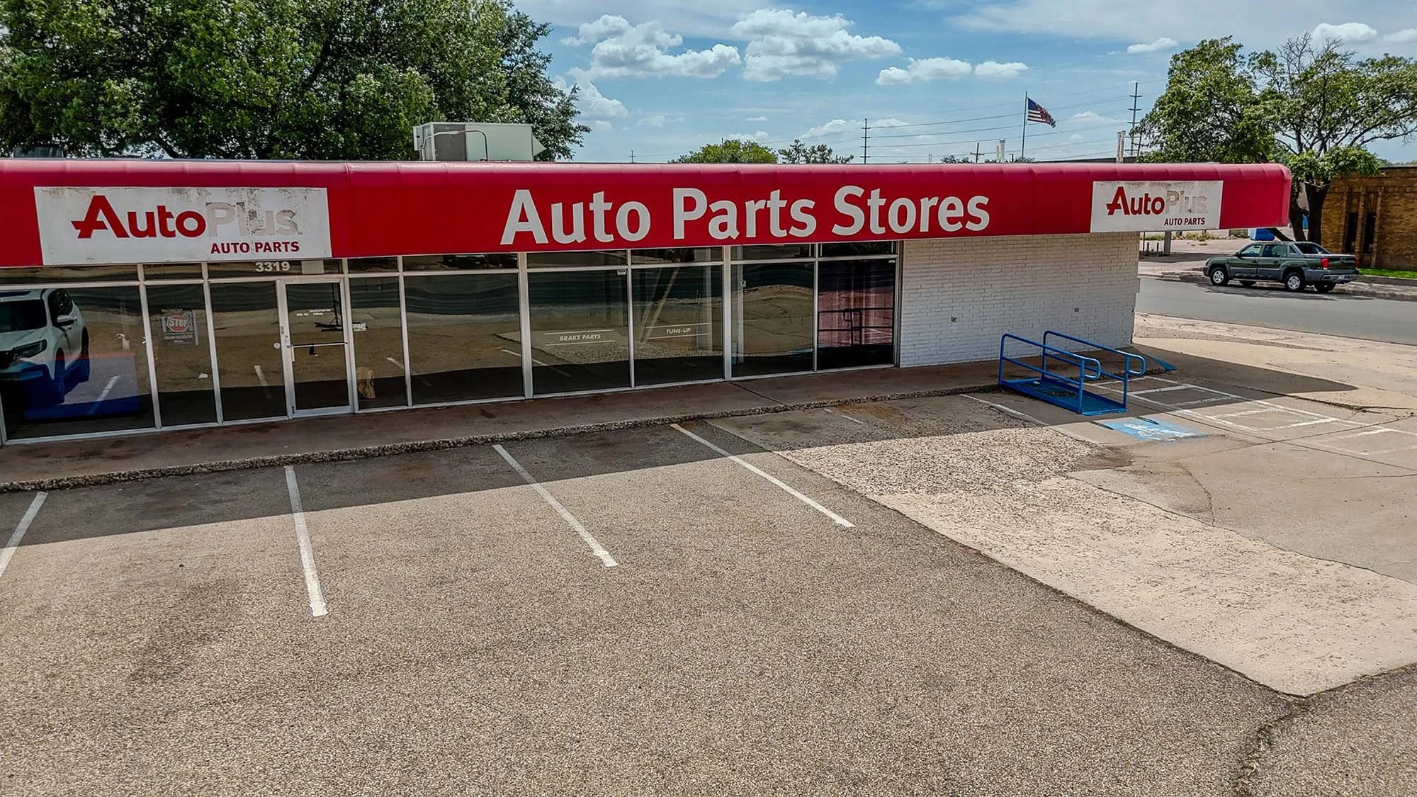 Exterior view of an Auto Parts Store with a red sign that reads 'Auto Parts Stores' and has the Auto Plus logo. There are empty parking spaces in front of the store, a blue wheelchair accessible ramp, and a parked SUV across the street. The building