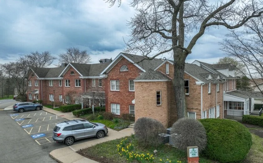 Apartment complex with brick buildings, parking lot with a few cars, surrounded by trees and bushes, on a cloudy day.