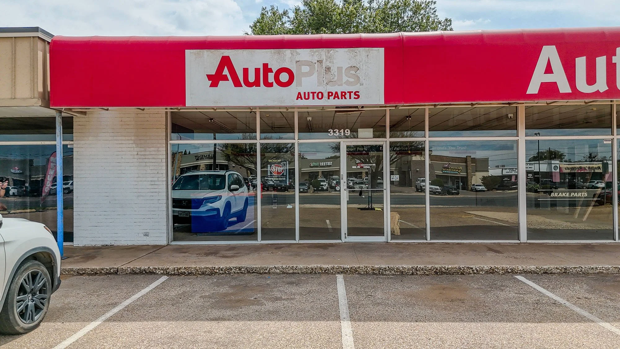 Auto Plus auto parts store storefront with glass windows, parking lot in front, and a red awning with the Auto Plus logo.