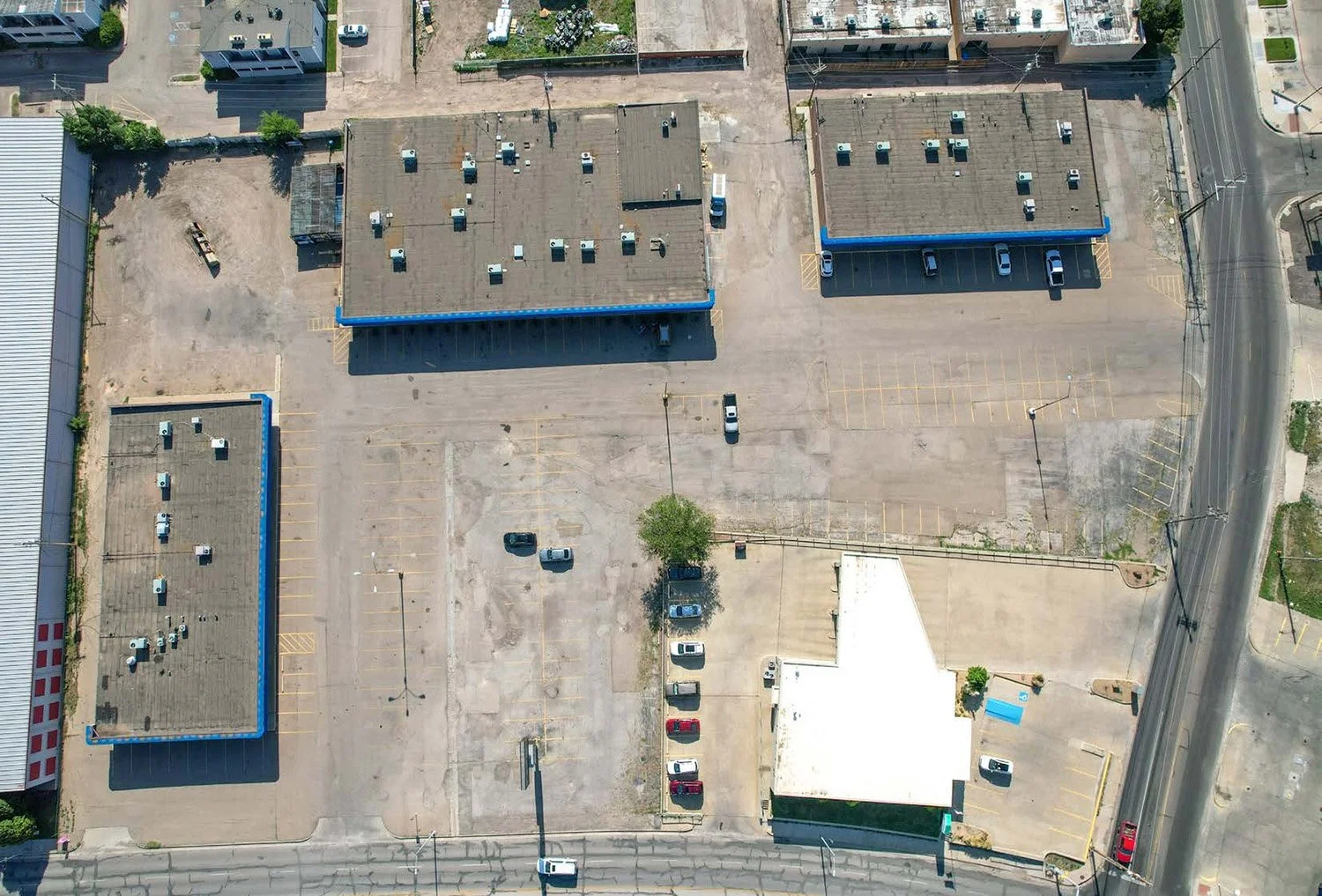 An aerial view of a parking lot with a few parked cars, surrounded by commercial buildings and streets, with some empty parking spaces and a few trees.