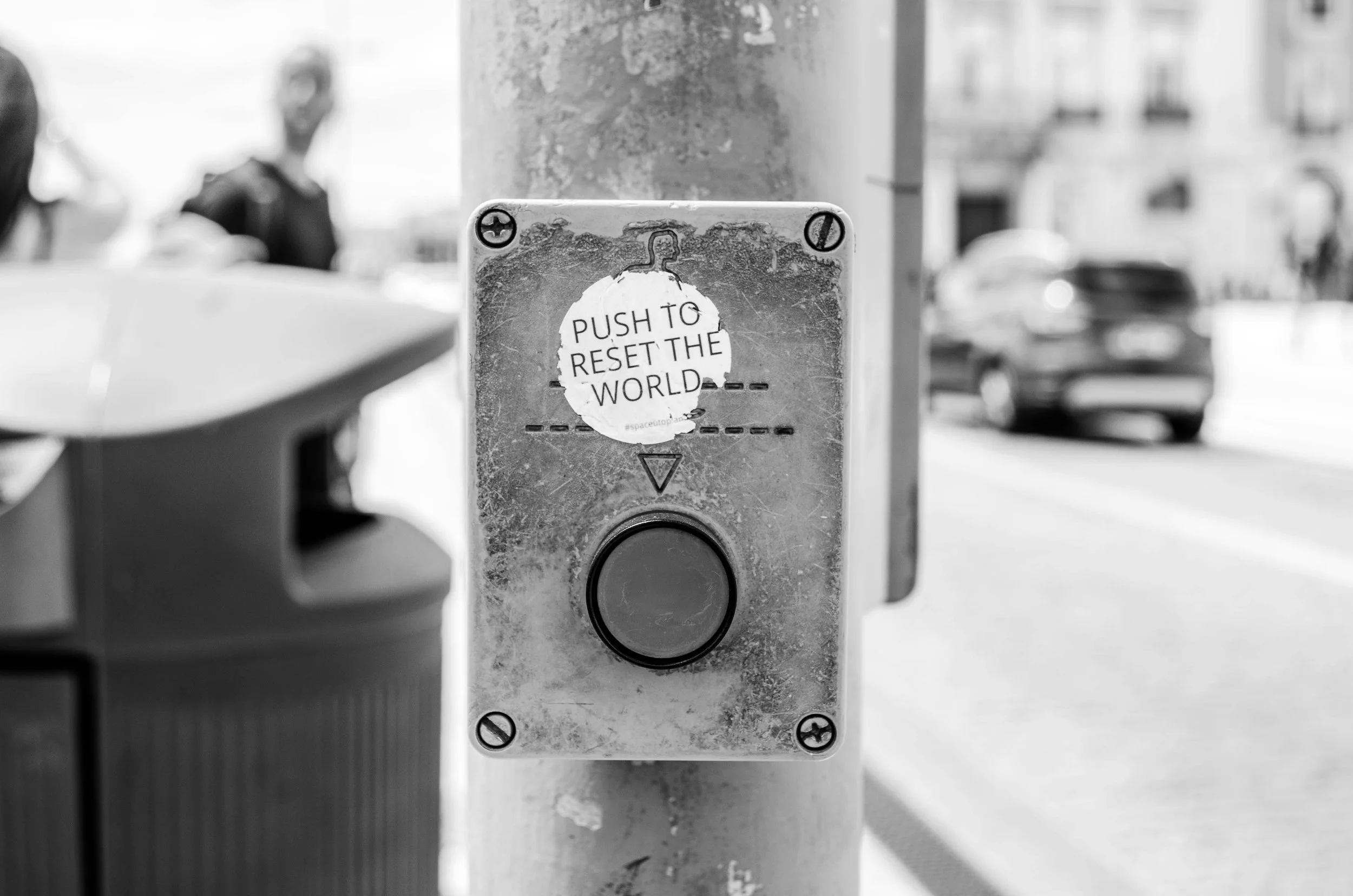 Close-up of a weathered button with a sticker that reads 'PUSH TO RESET THE WORLD,' on a city sidewalk, with blurred people and cars in the background. Used to symbolize Brainspotting as a reset for the nervous system.