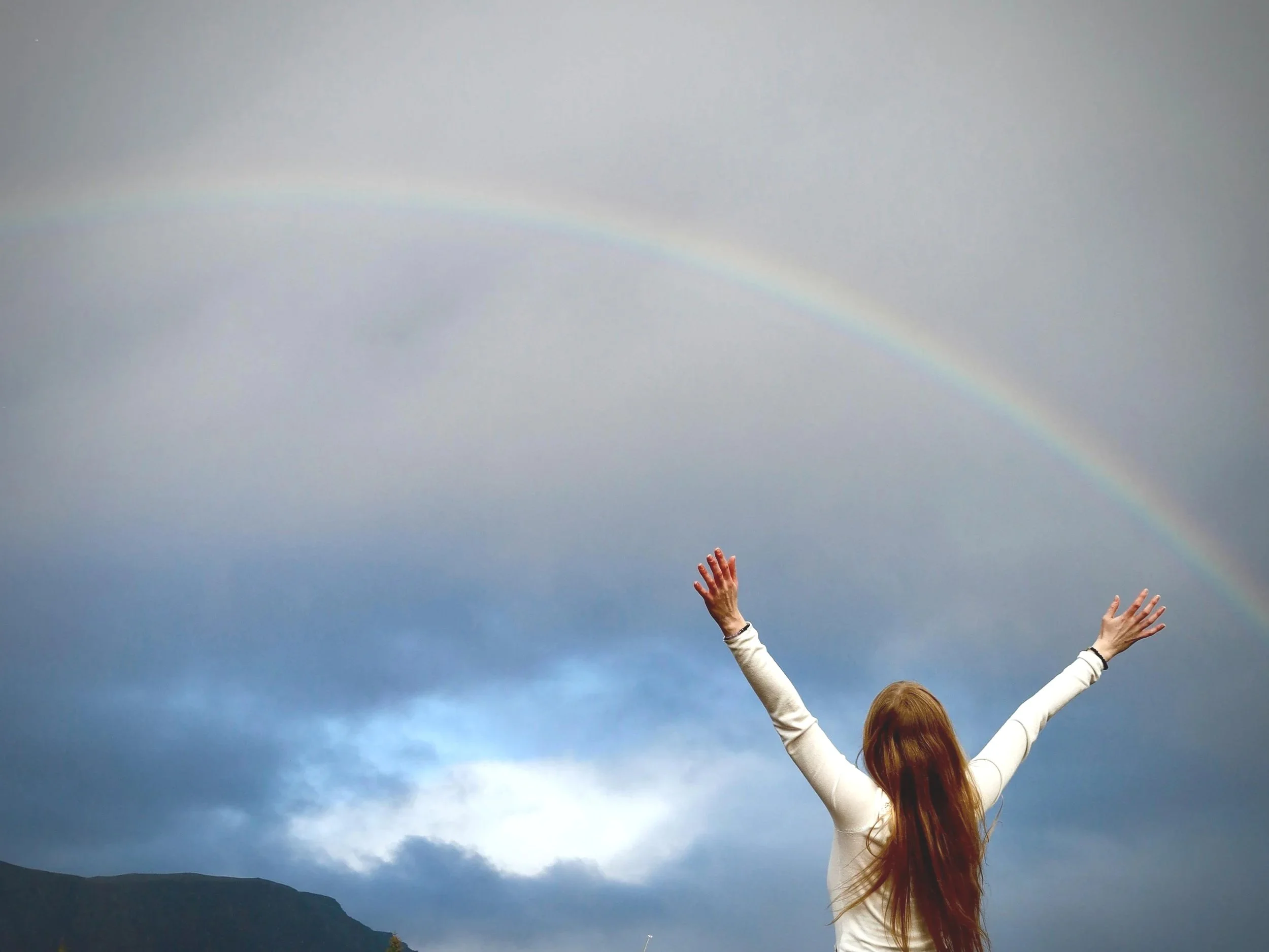 A woman with long red-brown hair wearing a white long-sleeve shirt stands outdoors with her arms raised, facing a rainbow against a cloudy sky. Displayed as a symbol of hope for grief and loss on therapy website.