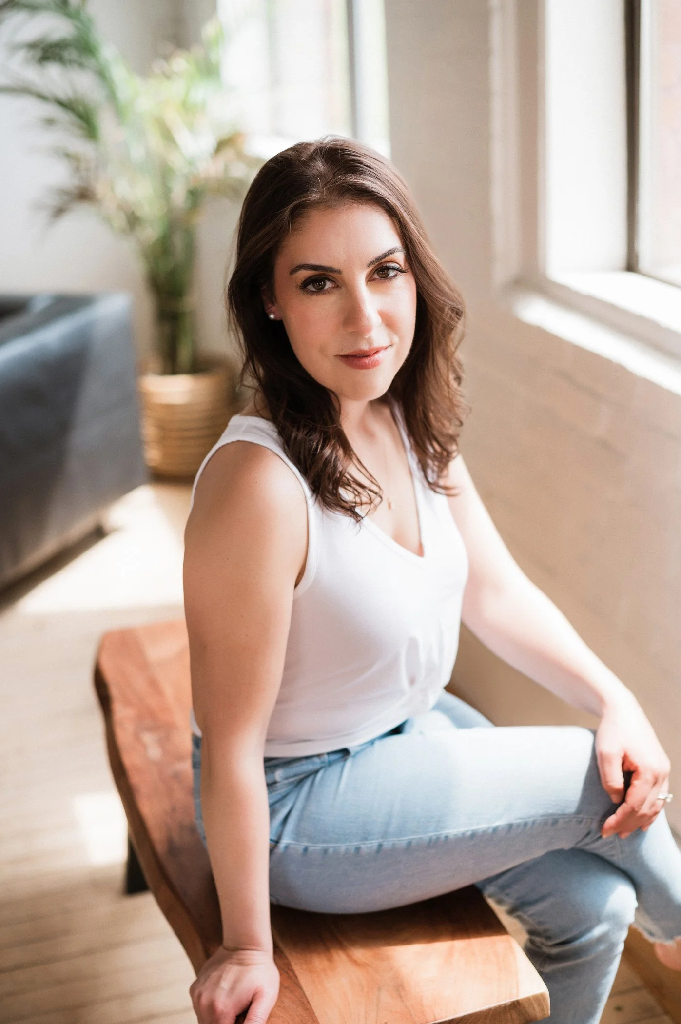 Therapist Rebecca Wilson, LCSW sitting on a wooden bench near a window, wearing a white top and light blue jeans, with green plants and a couch in the background.