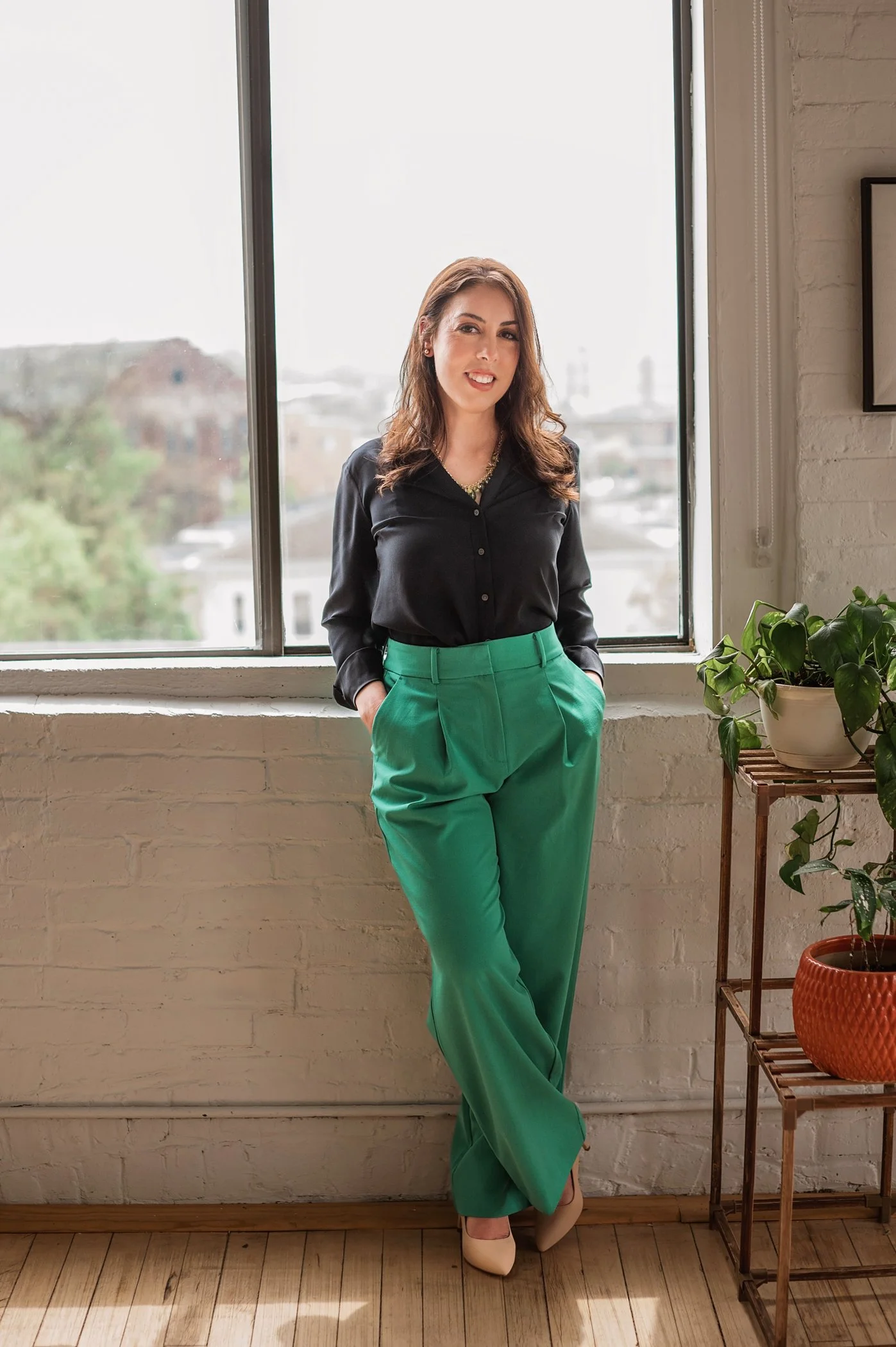 Therapist Rebecca Wilson, LCSW in Southington, CT smiling and leaning against wall in front of tall window next to green plants.