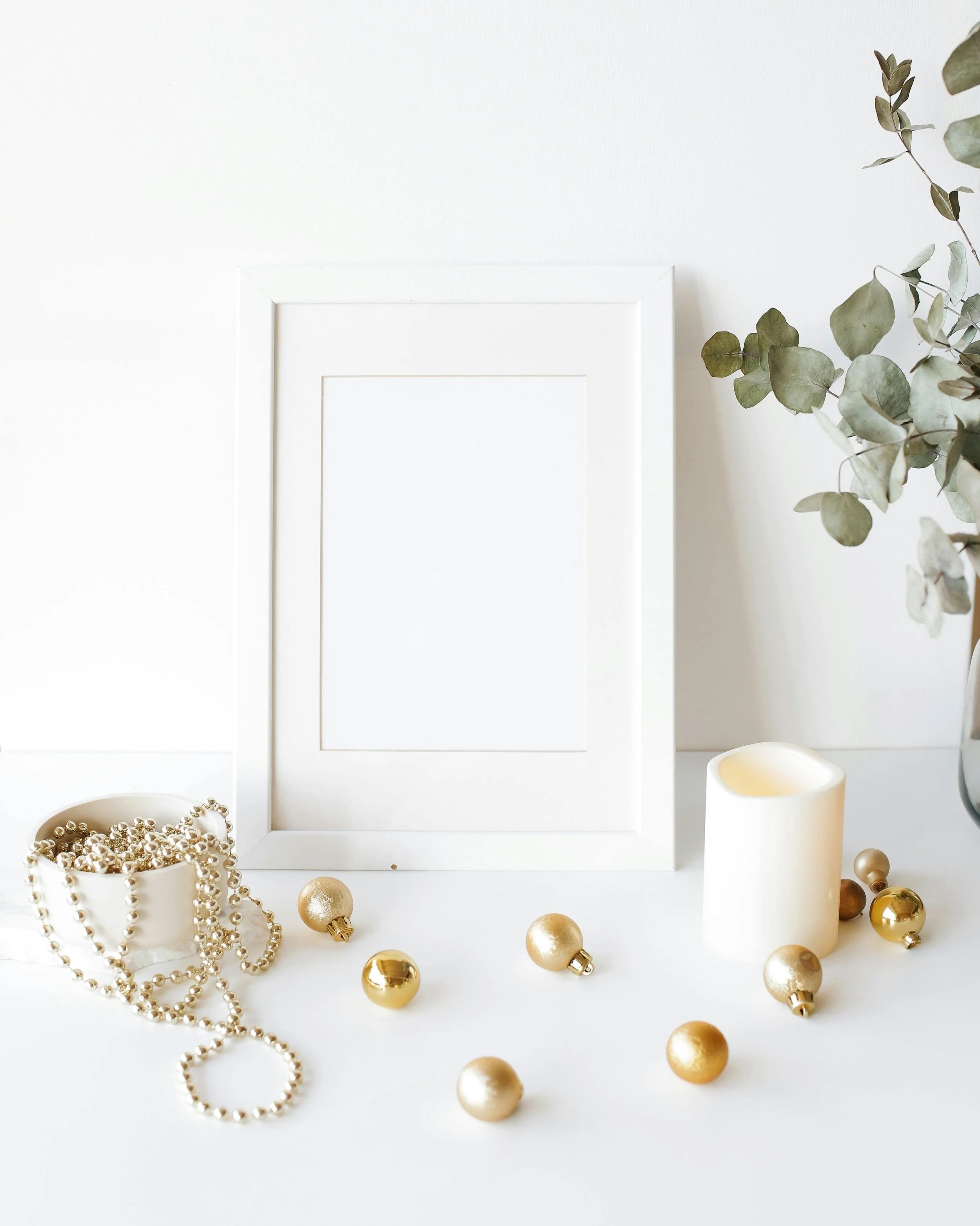 White picture frame, jar with gold beads and beads scattered across next to a glowing candle and eucalyptus.