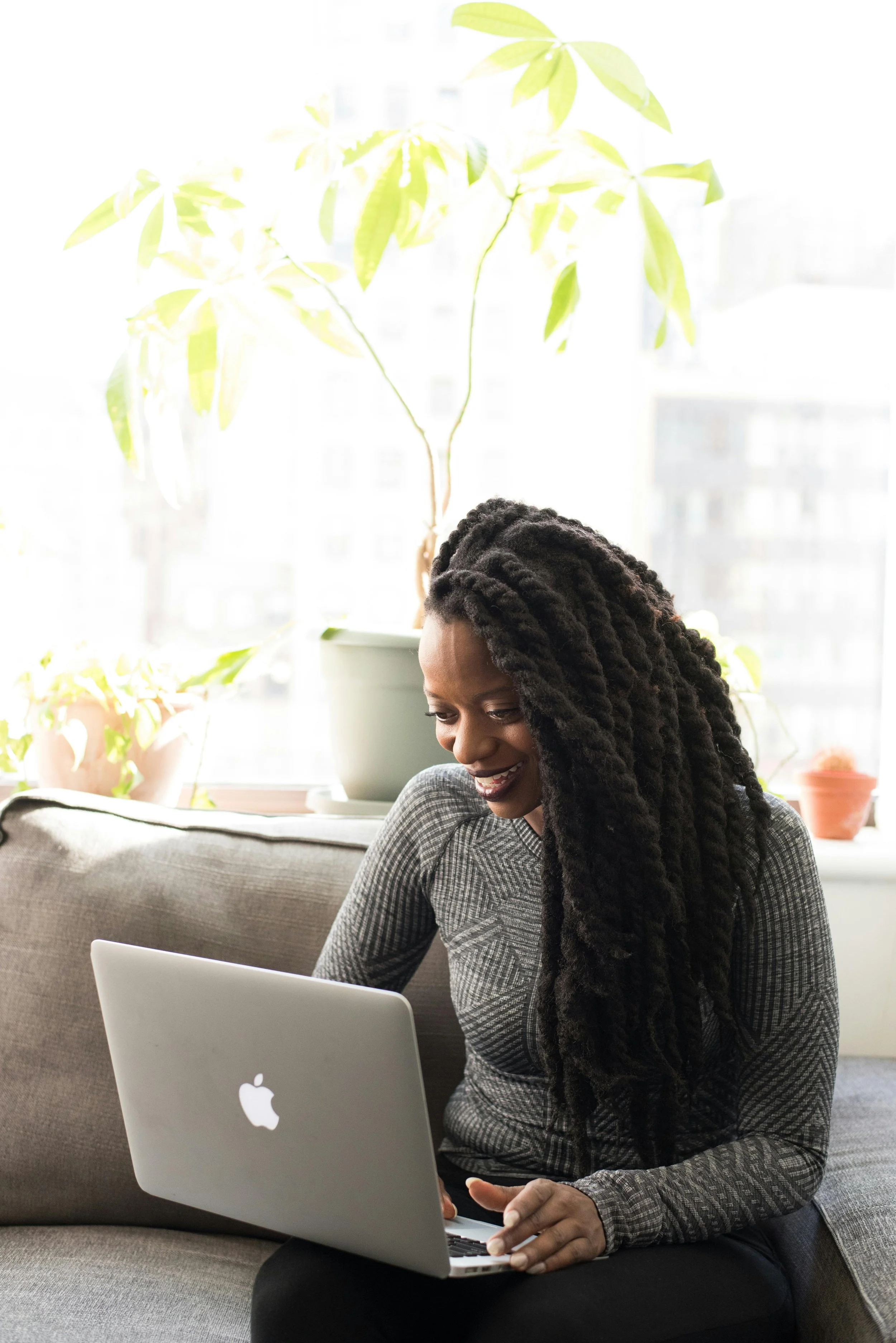 A woman with long, curly black hair smiling during an online therapy session on a  laptop, sitting on a gray couch in a bright room with potted plants on a window sill behind her.