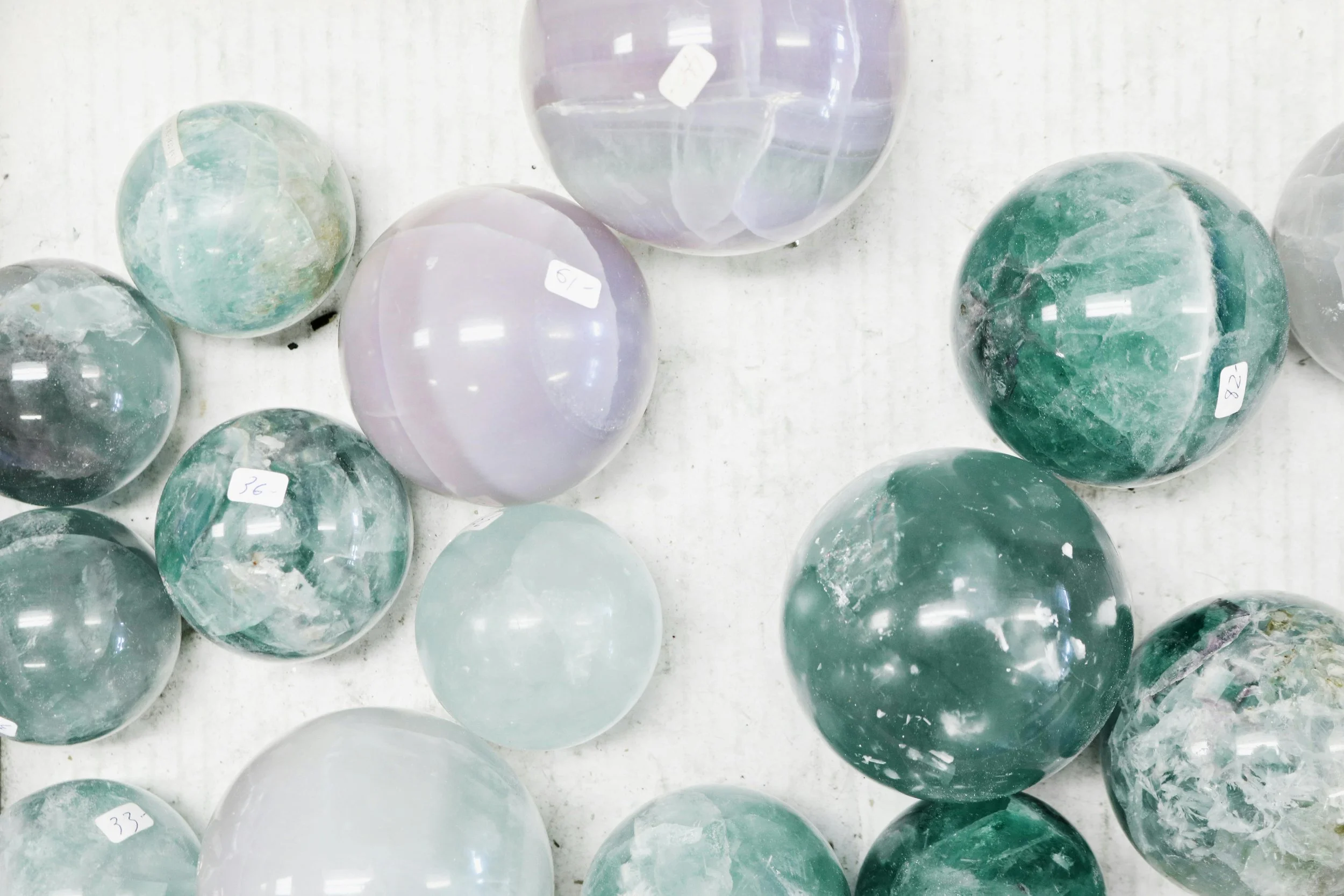 Collection of polished semi-precious stones and spheres in various shades of green, purple, and white, with some labeled with prices, on a white surface.