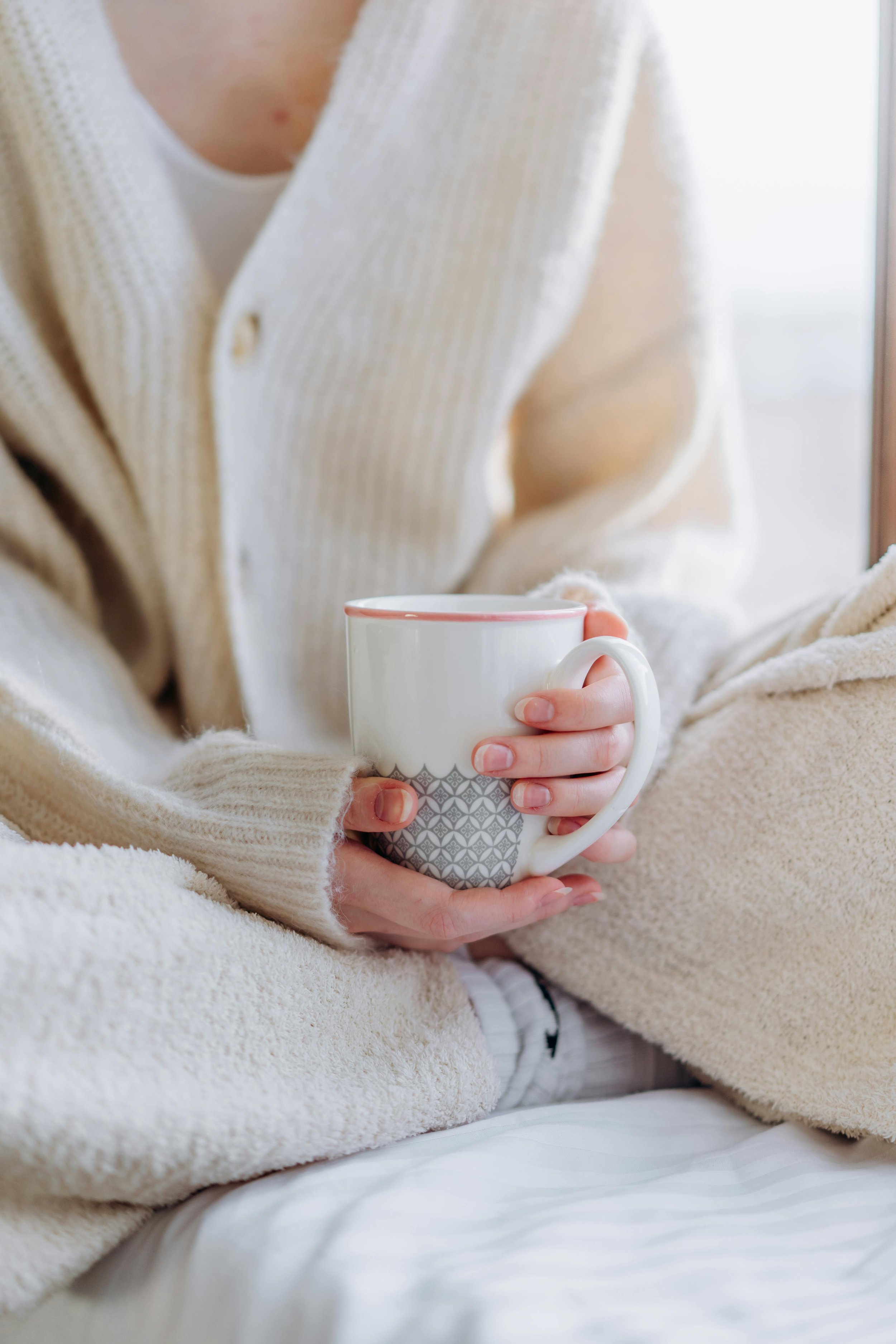 Person in cream-colored cardigan holding a gray and white mug, sitting on a bed with cream and beige blankets.