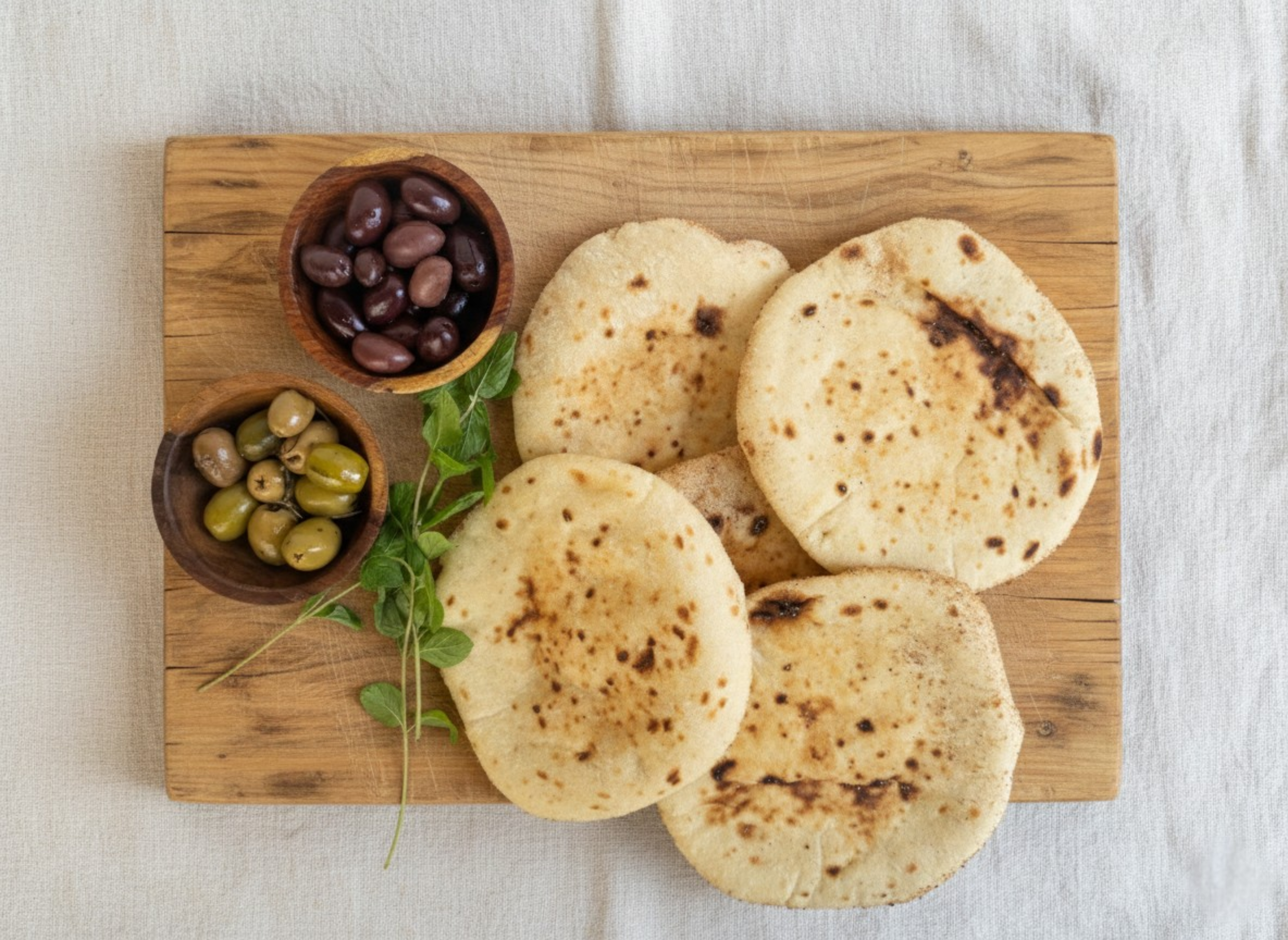 A wooden cutting board with four Egptian baladi breads, two small bowls of black and green olives, and some green herbs on a light fabric surface.