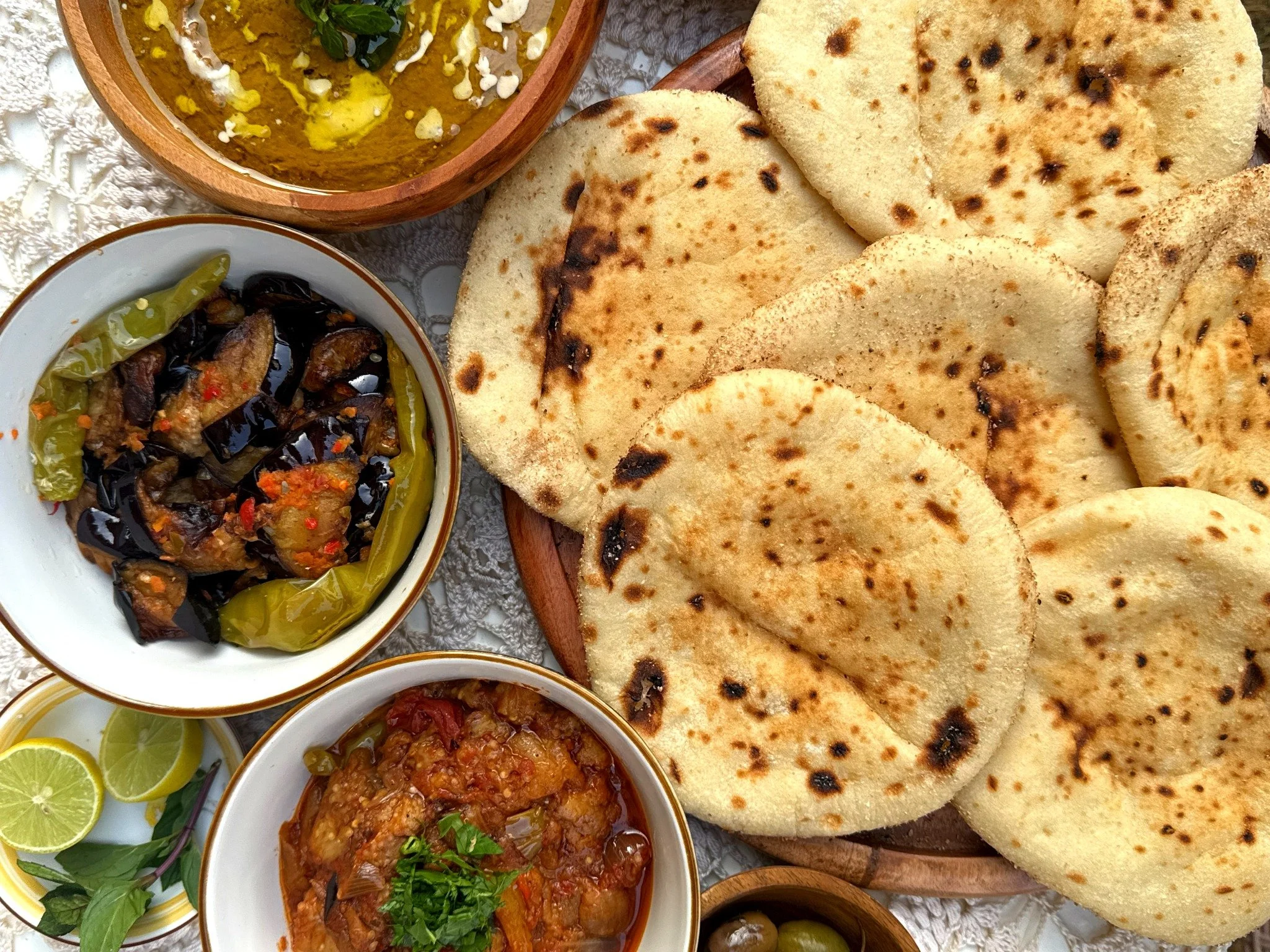 Plate of naan bread with bowls of vegetable curry, eggplant and green chili, and a side of lemon wedges and herbs.
