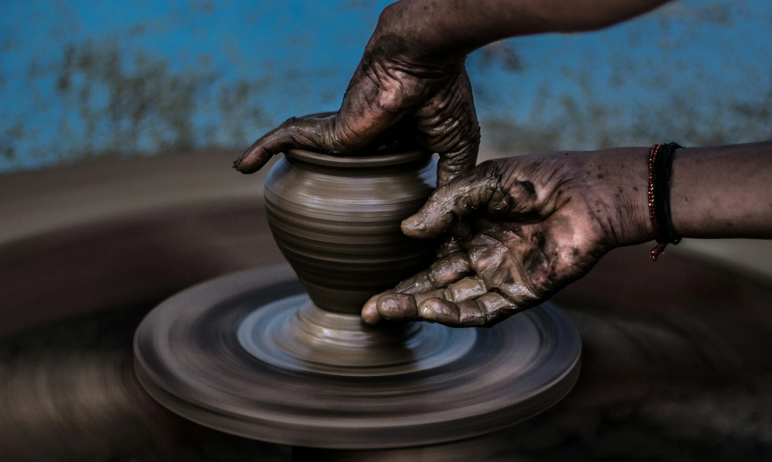 Hands of a person shaping pottery on a spinning wheel with clay. The hands are covered in wet clay, and the background is blurry.