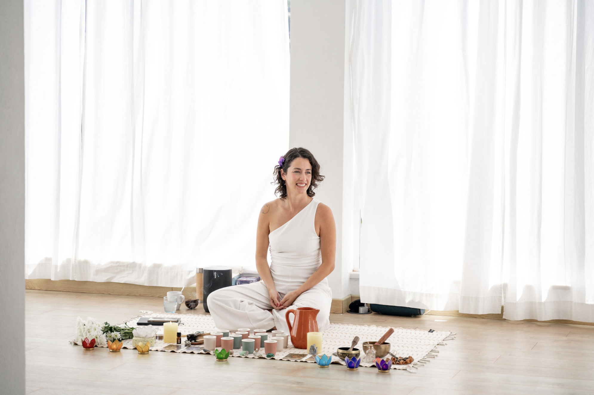 A woman sitting cross-legged on a rug surrounded by various colorful candles, bowls, and a large orange pitcher in a bright room with white curtains.