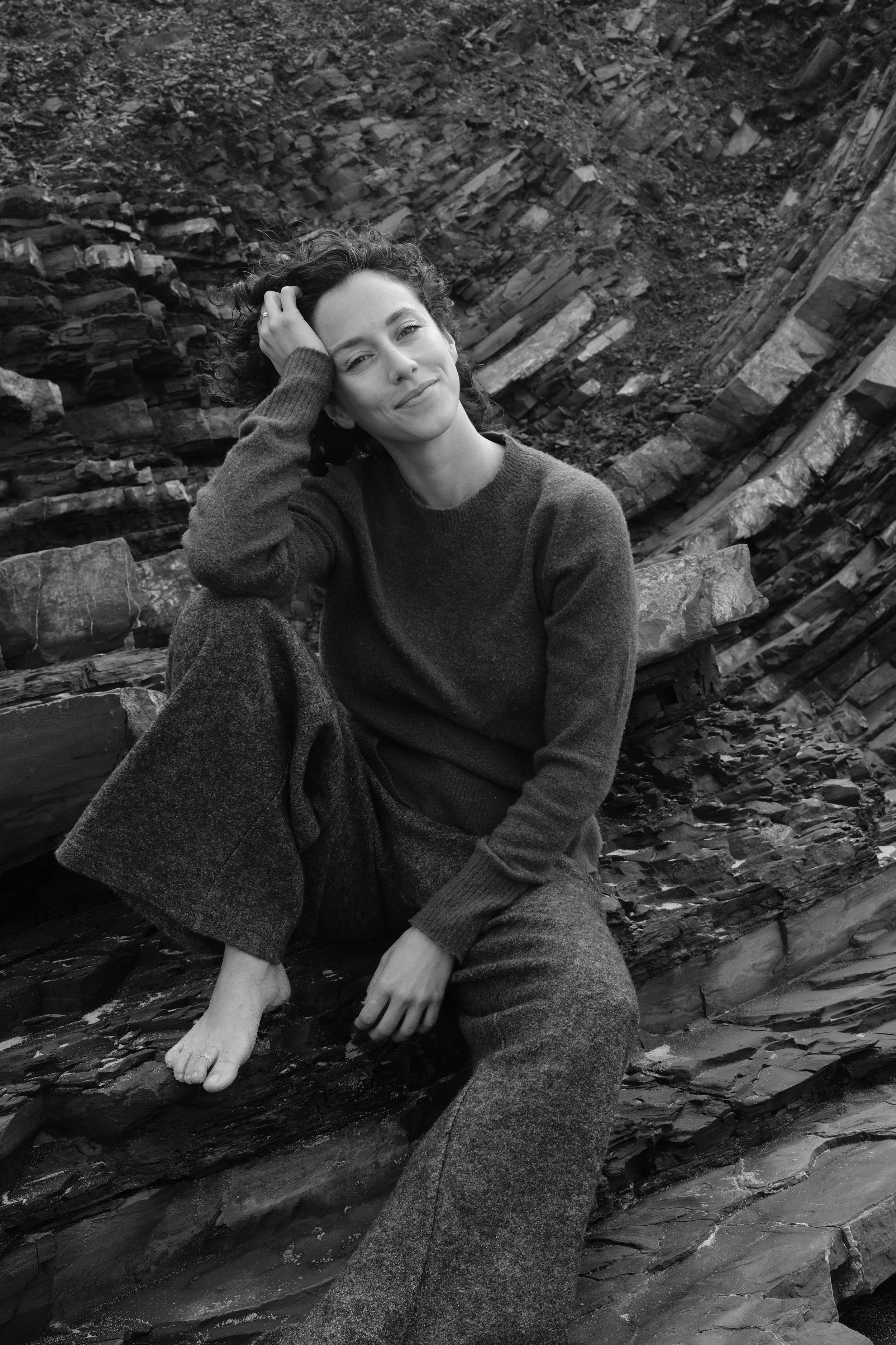 A woman sitting on layered rocks, dressed in a sweater and loose pants, smiling gently with one hand on her head in a natural outdoor setting.