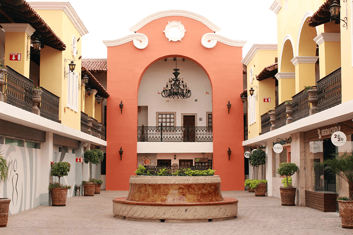 A colorful courtyard in Los Algodones, Mexico, features a round fountain, arched balconies, potted plants, and a grand chandelier.