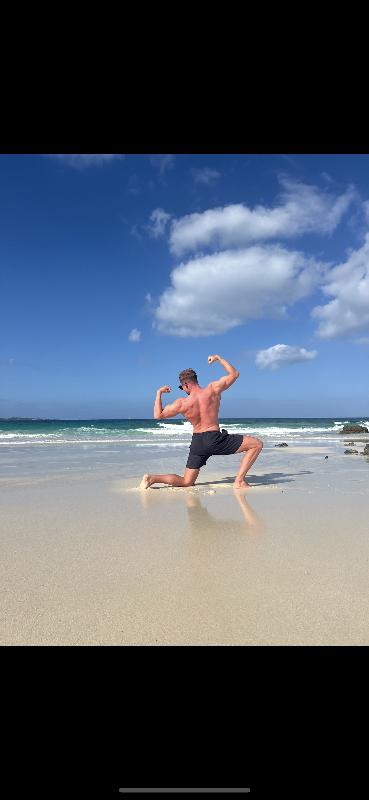 Mann in Badehose posiert als Bodybuilder am Strand unter blauer Himmel mit Wolken.