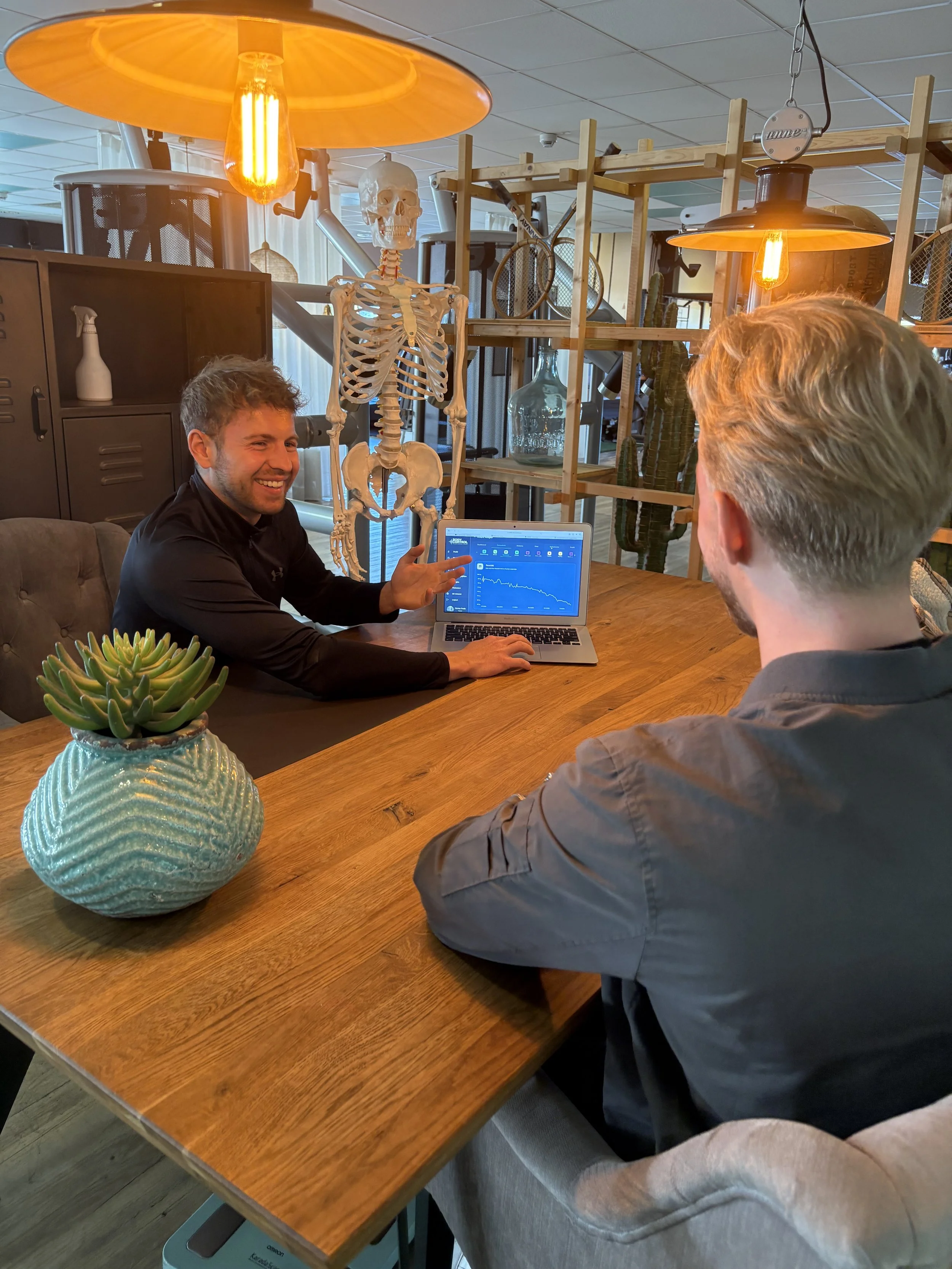 Zwei Männer sitzen an einem Holztisch in einem modernen Raum mit mehreren Lichtquellen, einem menschlichen Skelettmodell im Hintergrund, einer Pflanze in einer Vase auf dem Tisch und einem Laptop, der die Graphen für eine Datenanalyse zeigt.