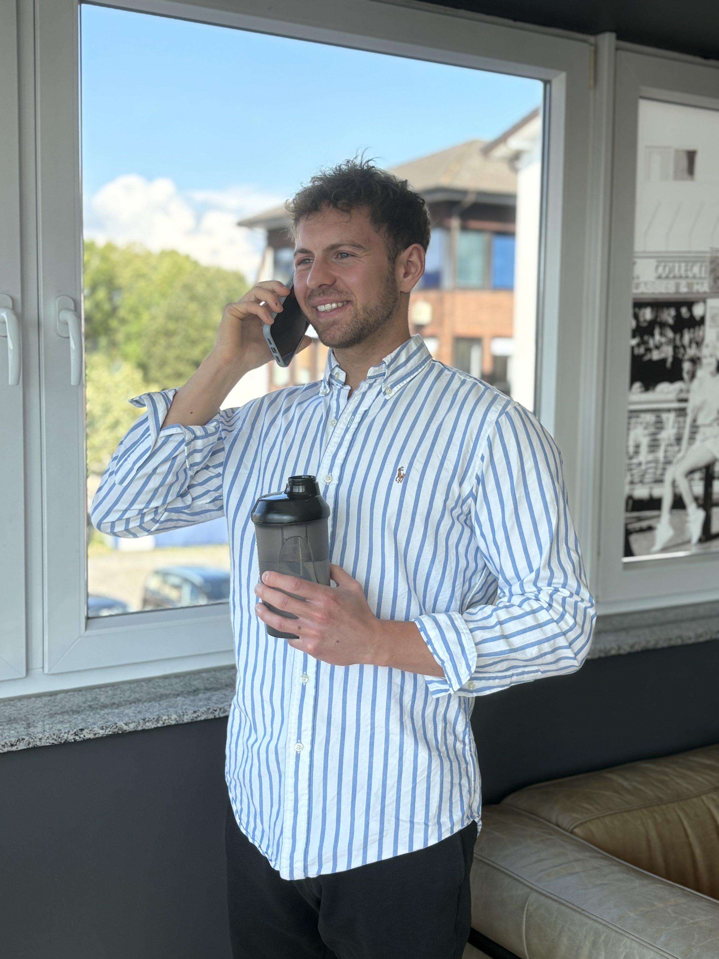 Junger Mann spricht im Büro am Telefon und hält eine Wasserflasche in der Hand, vor einem Fenster mit Blick nach draußen.