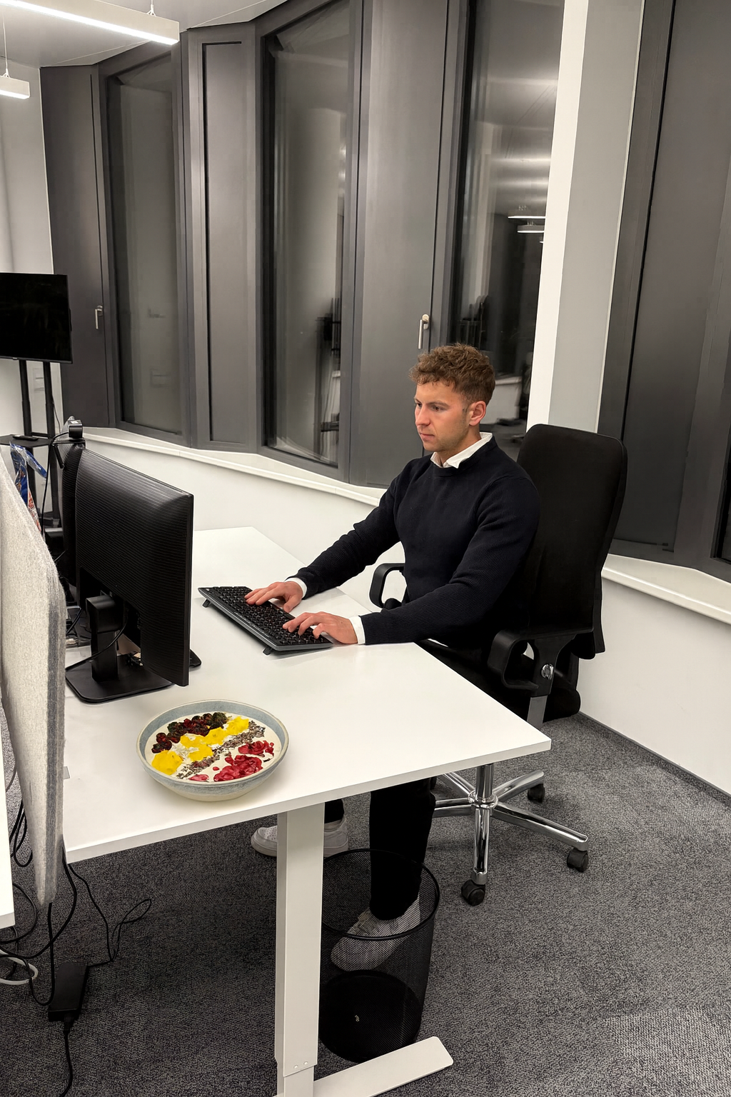 Ein Mann arbeitet an einem Schreibtisch im Büro vor einem Computer Monitor, während auf dem Tisch eine Schüssel mit verschiedenen Lebensmitteln steht.