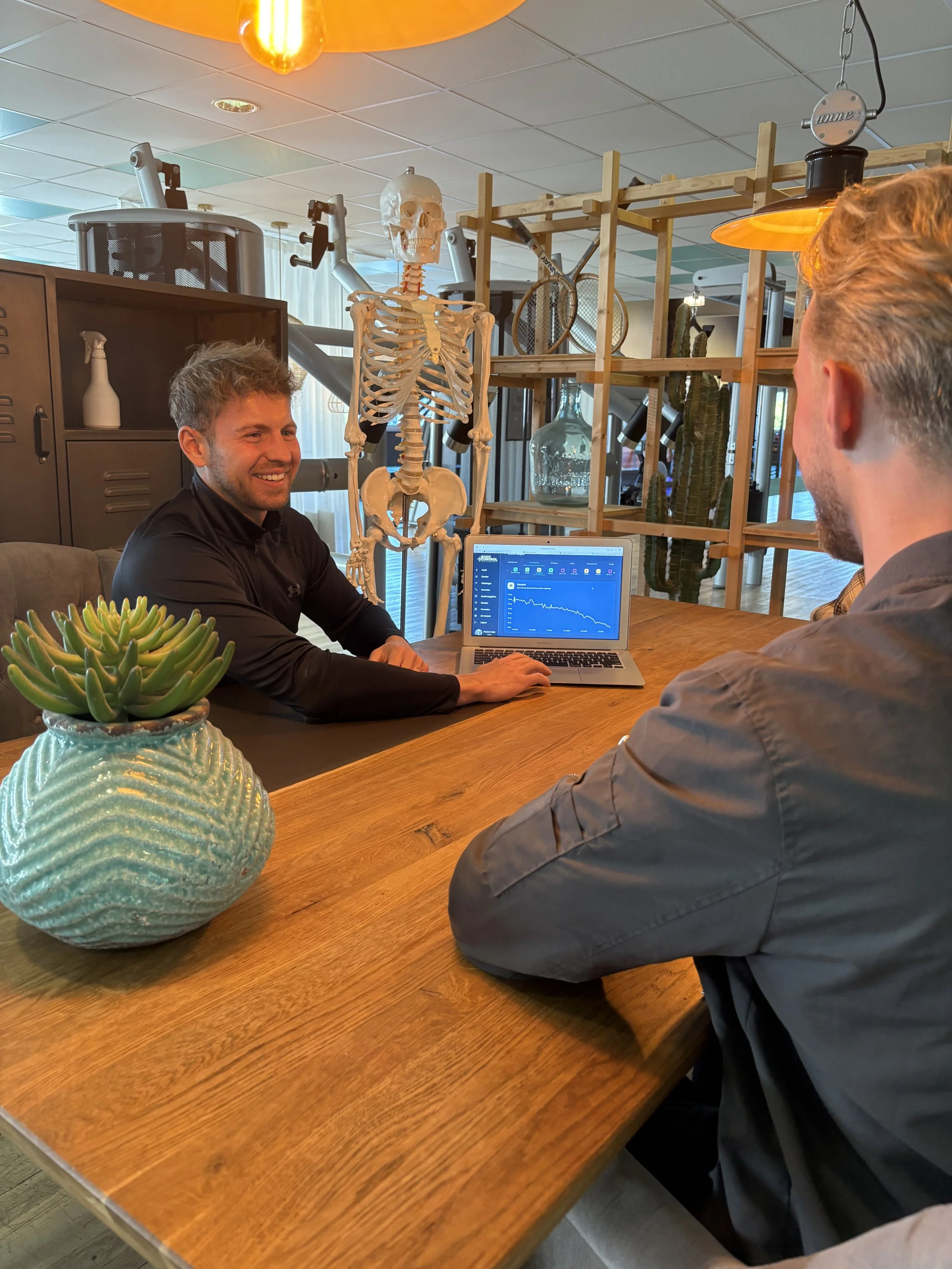 Zwei Männer unterhalten sich an einem Holztisch in einem Raum mit medizinischen oder wissenschaftlichen Elementen, darunter ein menschliches Skelettmodell und medizinische Geräte im Hintergrund.