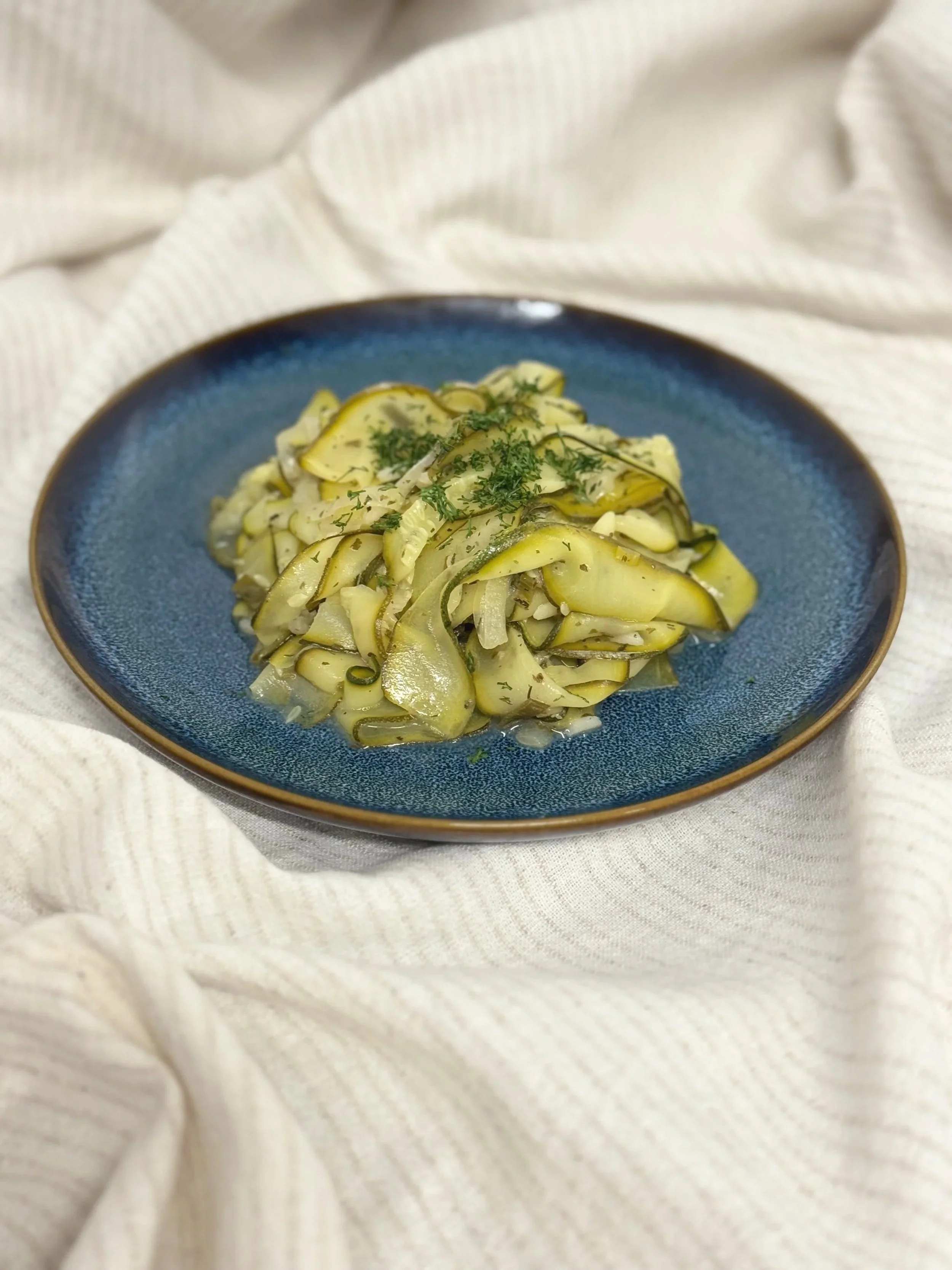 A blue plate with sliced zucchini and herbs, garnished with chopped herbs on a cream-colored cloth.