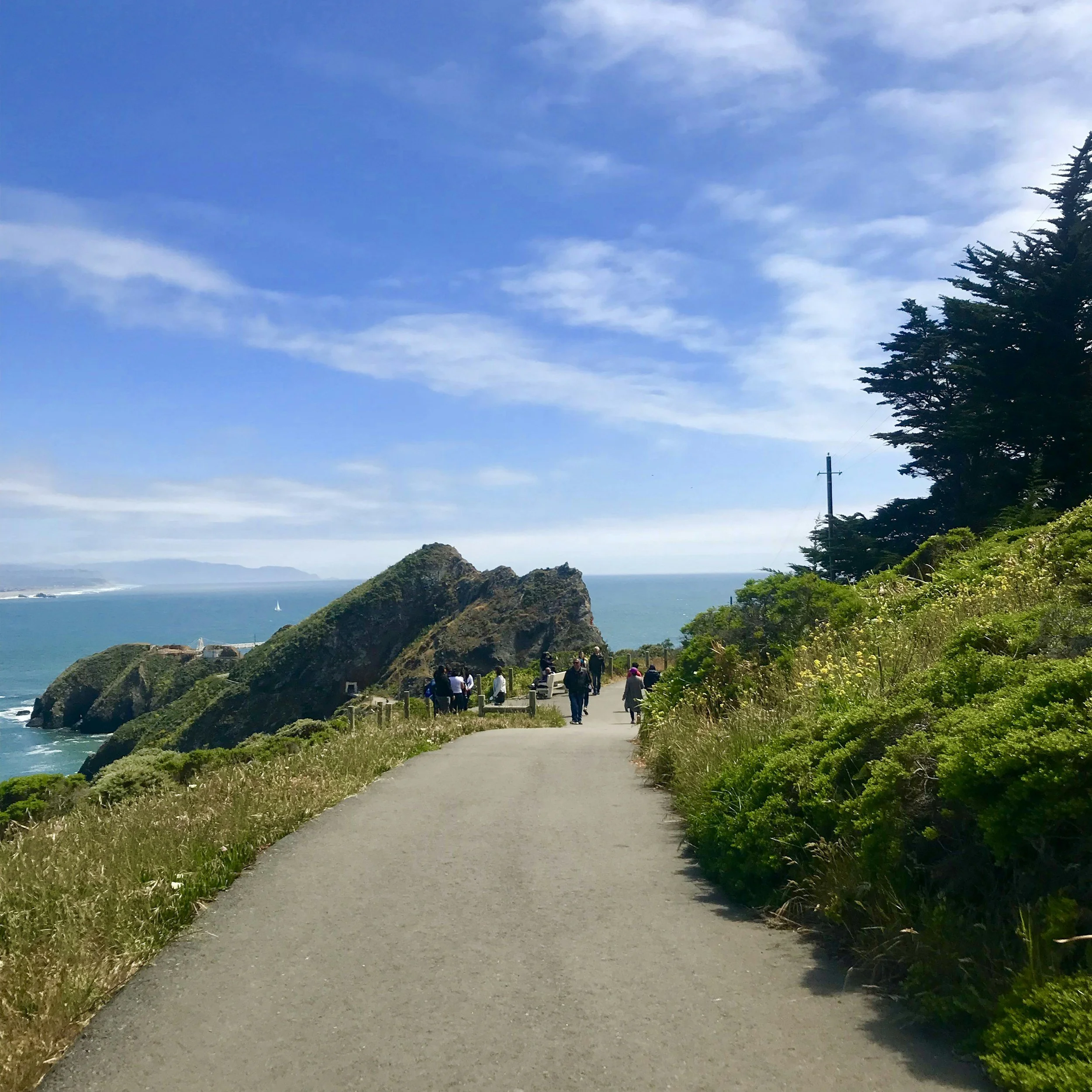 Trail along the coast with people walking, large rocks, greenery, blue sky with clouds, and ocean in the background.