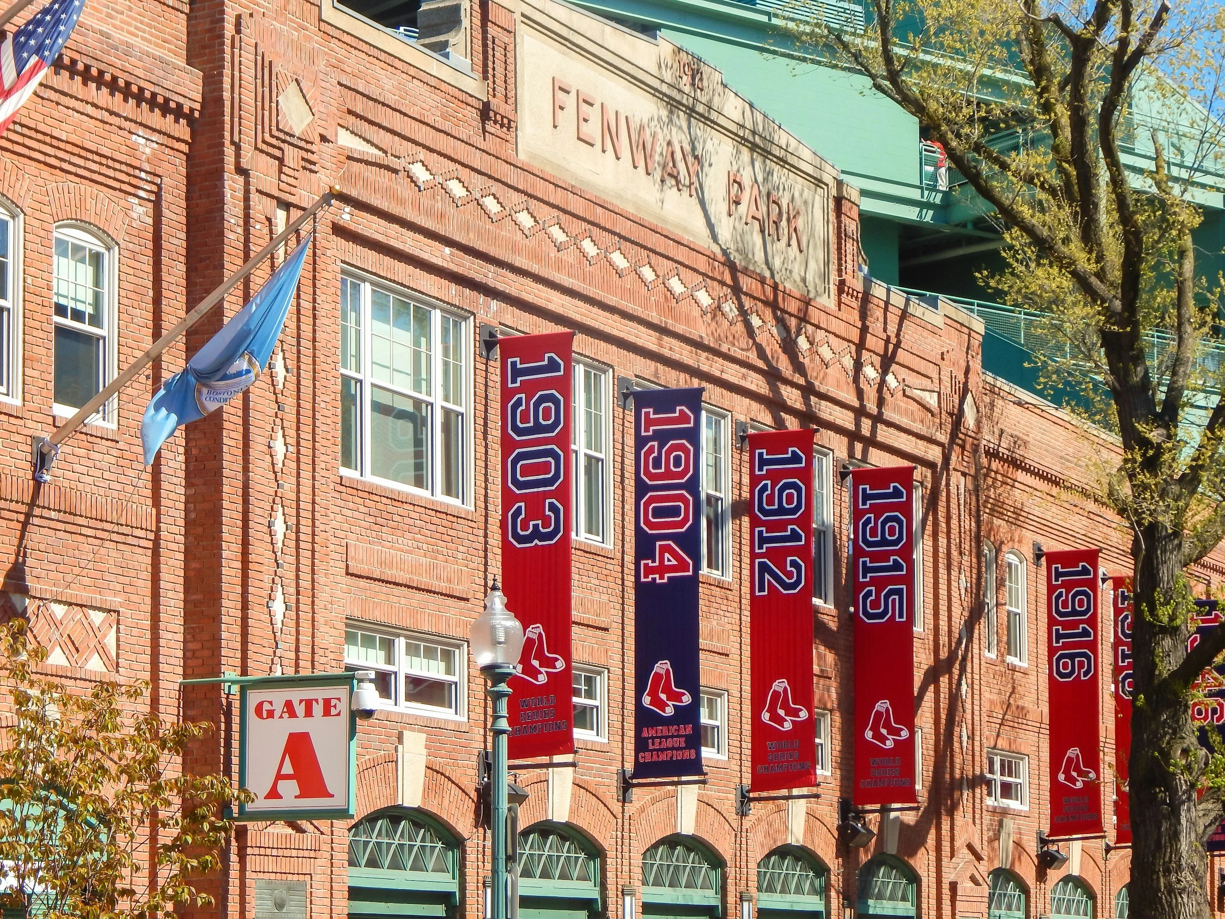 Exterior of Fenway Park with red banners displaying 2004, 2007, 2013, 2018, and 2019 World Series Champions, flags, and a gate sign labeled GATE A. A large green tree and parts of the stadium structure are also visible.
