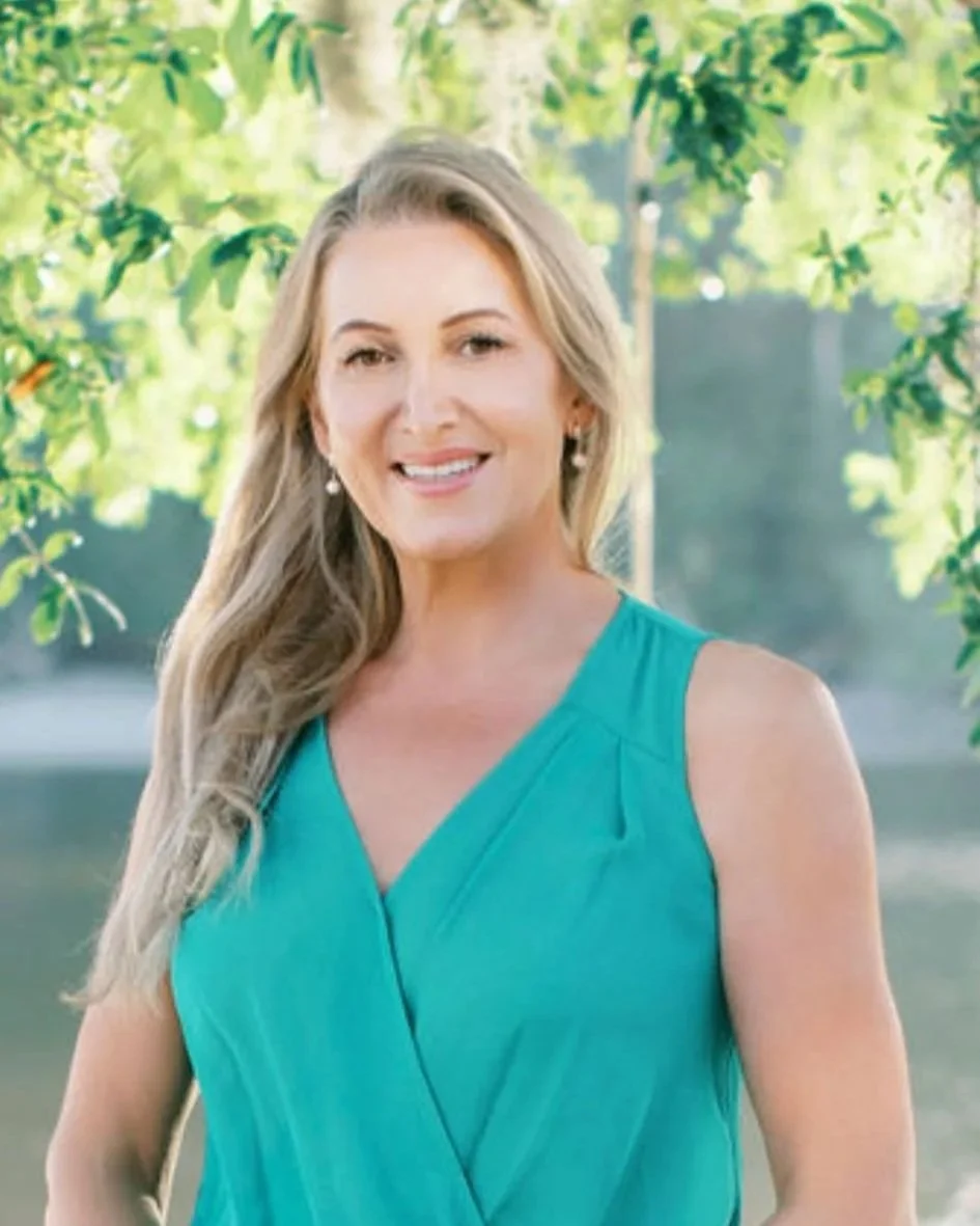 A woman with long blonde hair wearing a sleeveless teal dress, standing outdoors with green trees in the background, smiling at the camera.