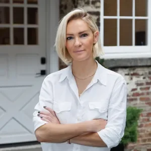 A woman with blonde hair in a white shirt standing outside a house with a white door and brick wall.