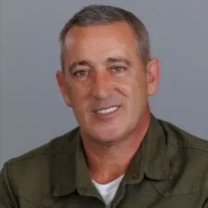 A man with short gray hair smiling, wearing a green jacket and white shirt, against a plain gray background.