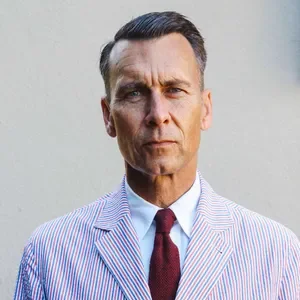 A man in a striped suit with a stern expression standing outdoors against a plain background.