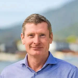 A man with short light brown hair wearing a blue collared shirt, standing outdoors with a blurred mountain and cloudy sky background.