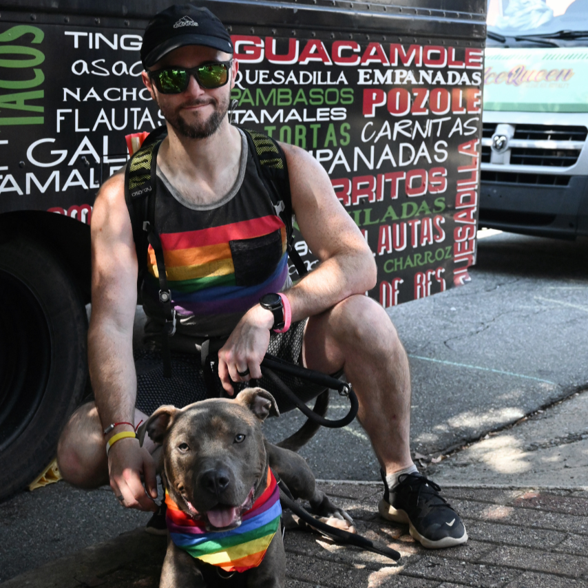 A man wearing sunglasses, a cap, and a rainbow tank top poses with a dog that is also wearing a rainbow bandana, at a street food stand with a menu in the background.