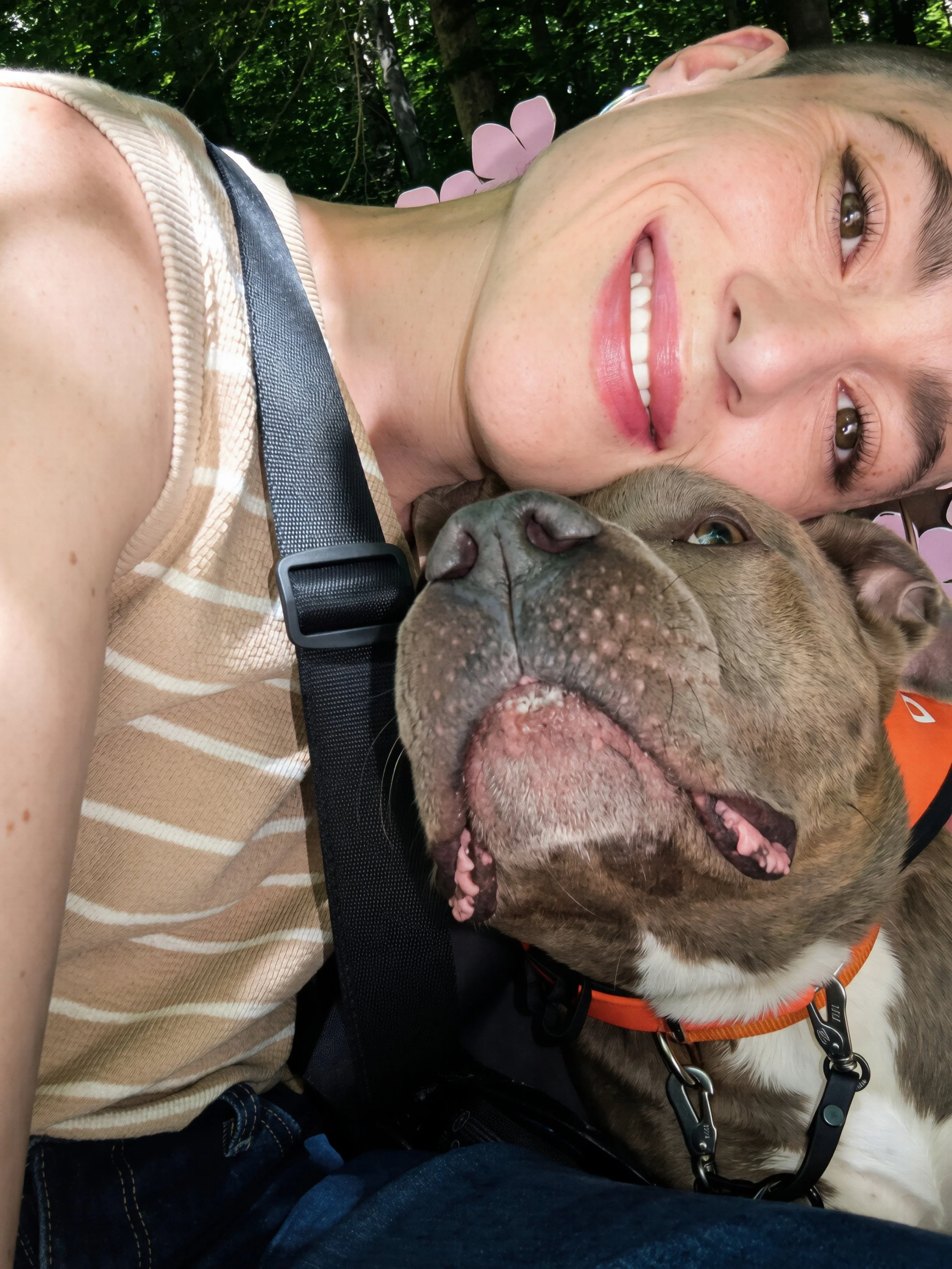Close-up of a smiling woman with brown hair lying next to a large, happy bulldog with its tongue out, outdoors with green trees in the background.