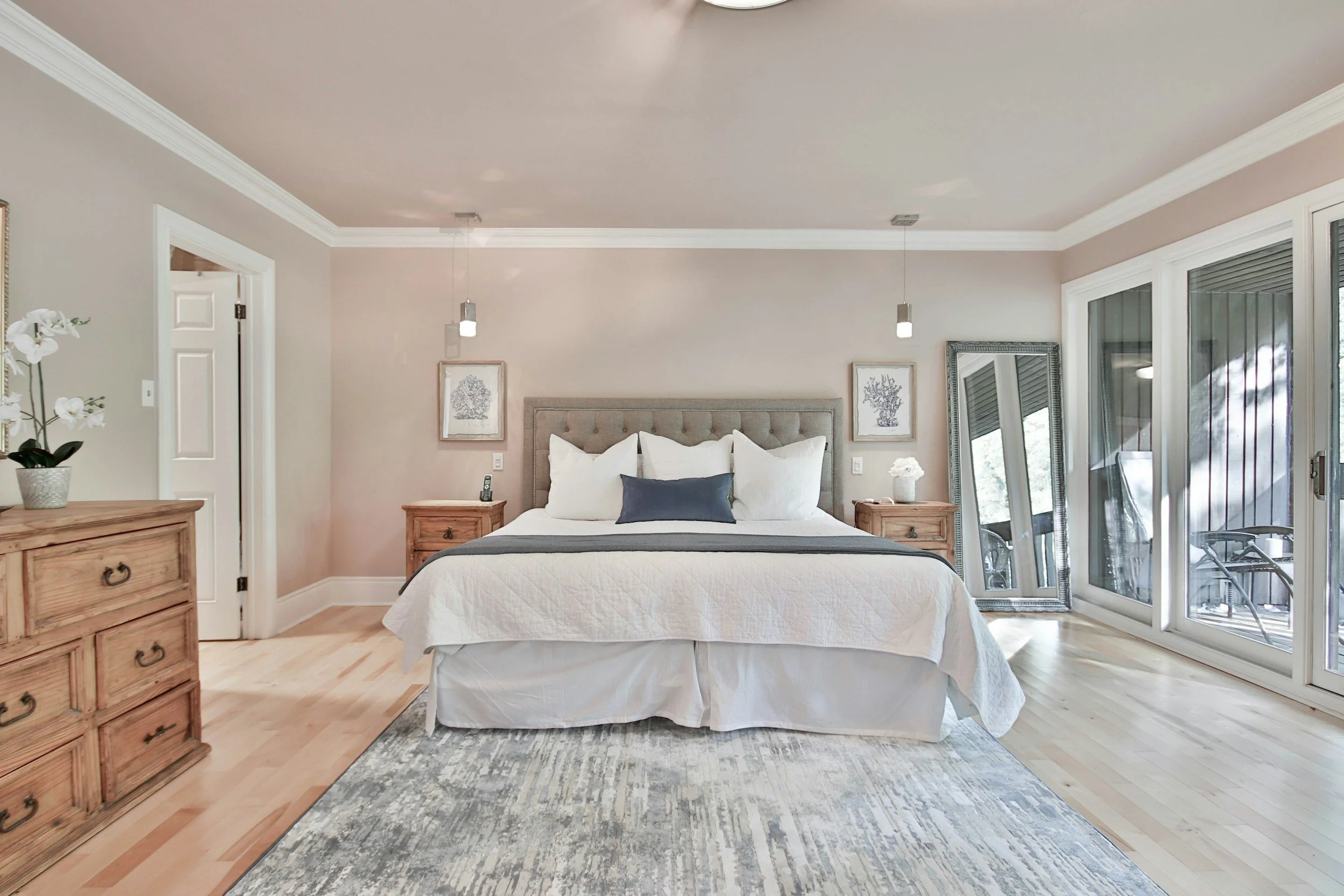 A bedroom with a bed, two wooden nightstands, a dresser, a large mirror, and sliding glass doors leading to a balcony, decorated in neutral tones.