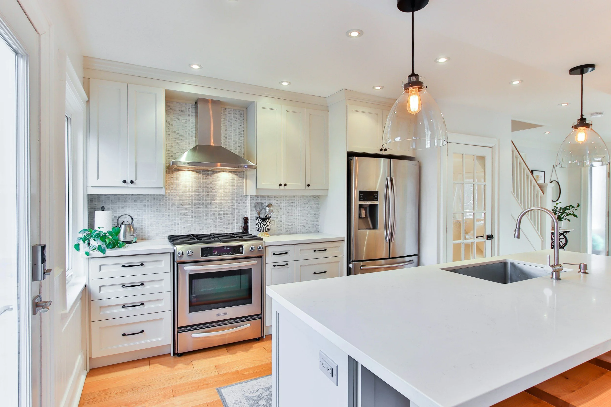 Modern kitchen with white cabinets, stainless steel refrigerator and oven, glass pendant lights, and a white island countertop with a sink.