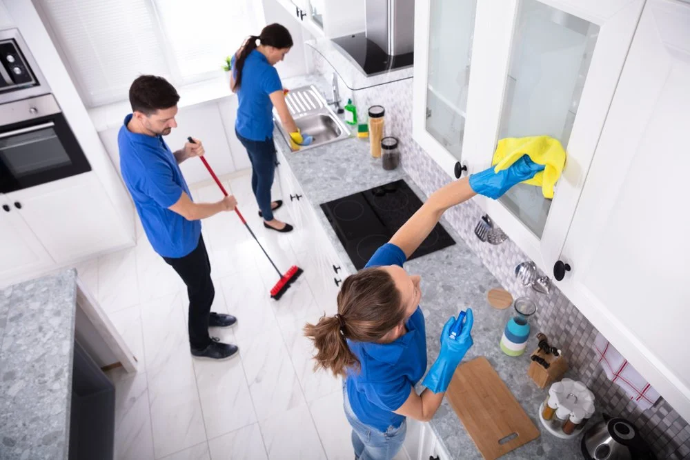 Three people cleaning the kitchen: a woman wiping the cabinet with a yellow cloth and wearing yellow gloves, a man sweeping the floor with a broom, and another woman at the sink washing dishes or cleaning.