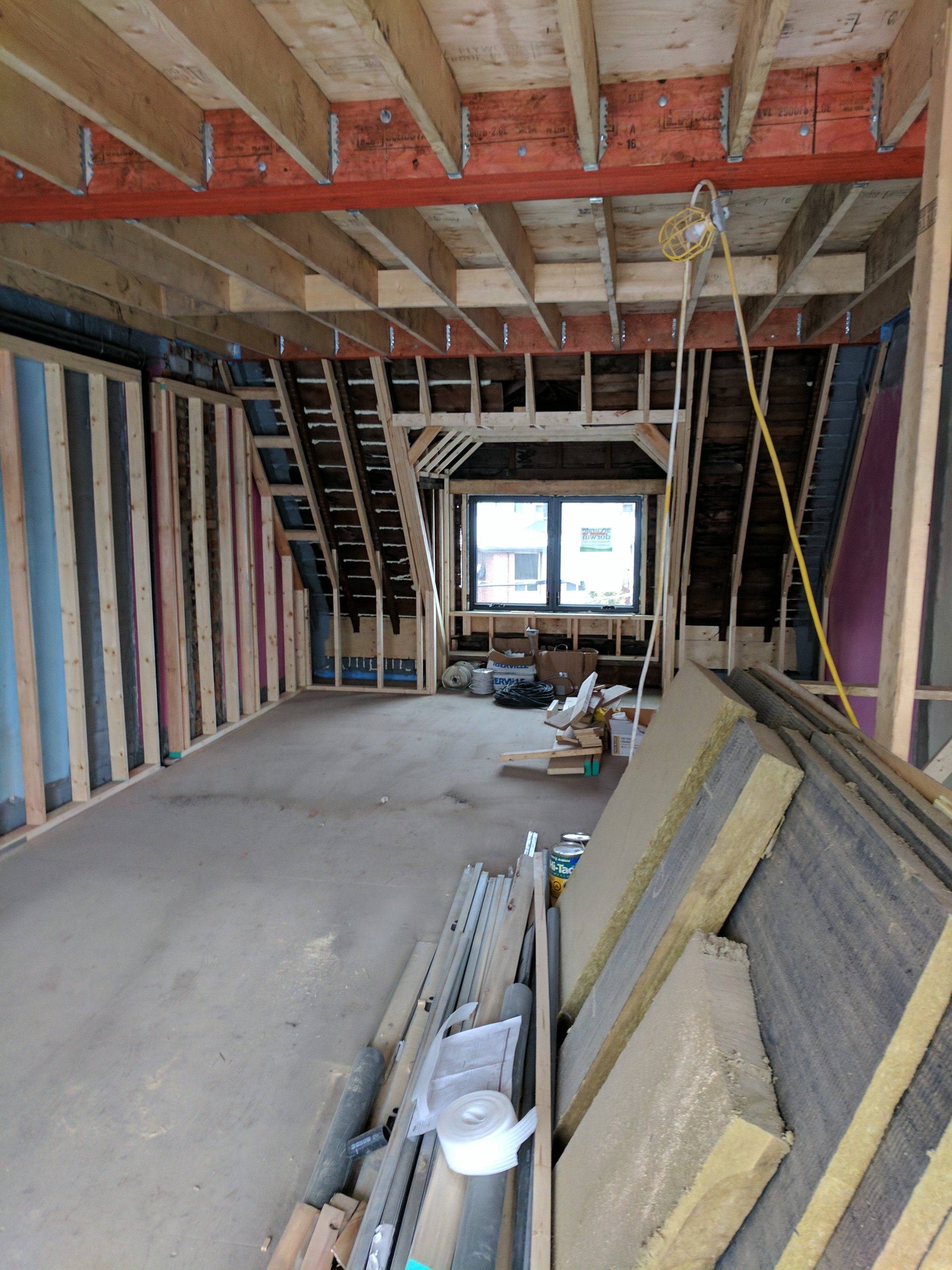 Interior view of an unfinished attic with exposed wooden framing, insulation materials, construction tools, and supplies scattered on the floor, with a window at the far end.