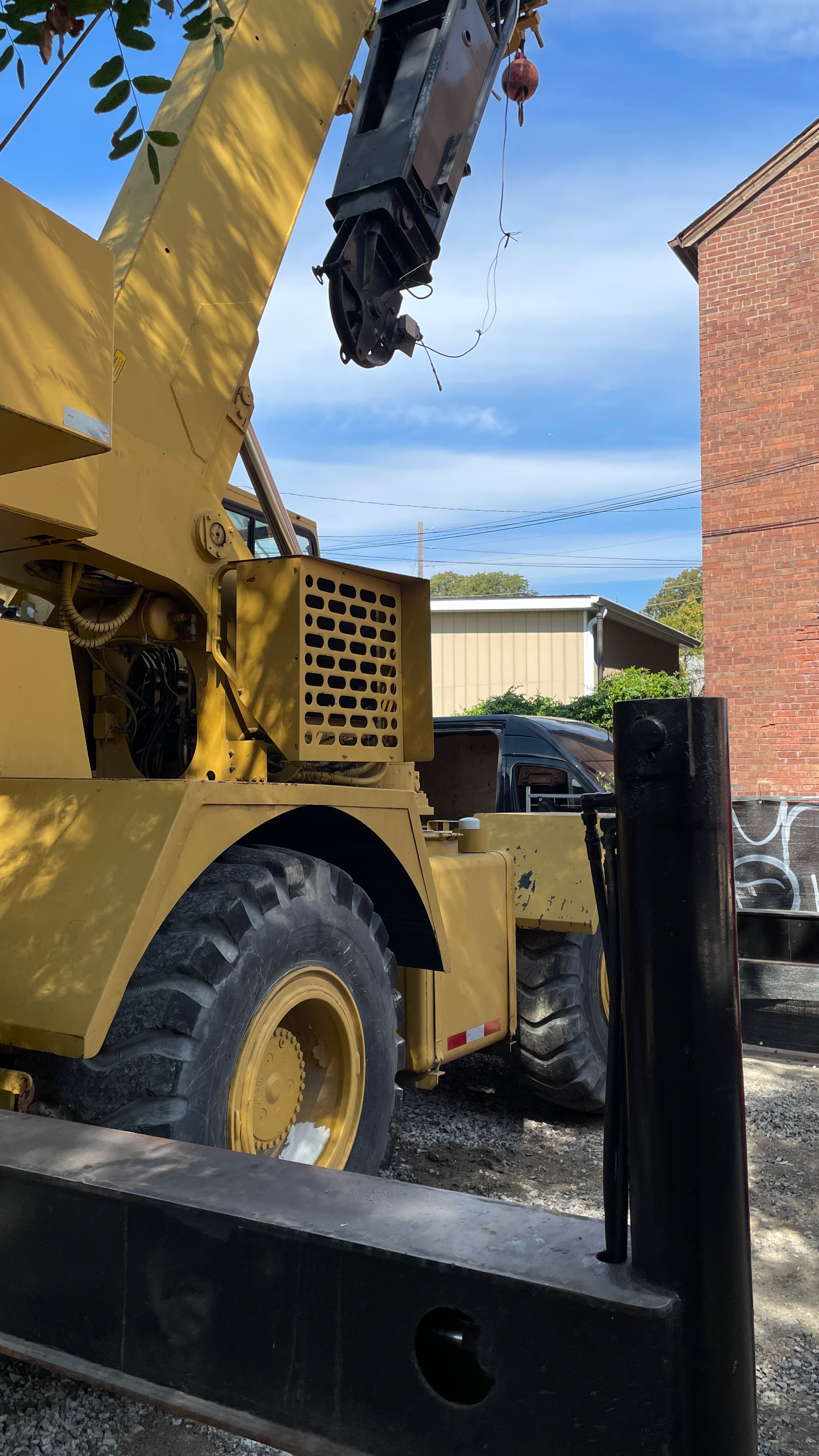 Close-up of a yellow construction crane with a black hook hanging from an extended arm, parked on a gravel lot near a black truck and a brick building with a concrete barrier in the foreground and a blue sky with some clouds in the background.