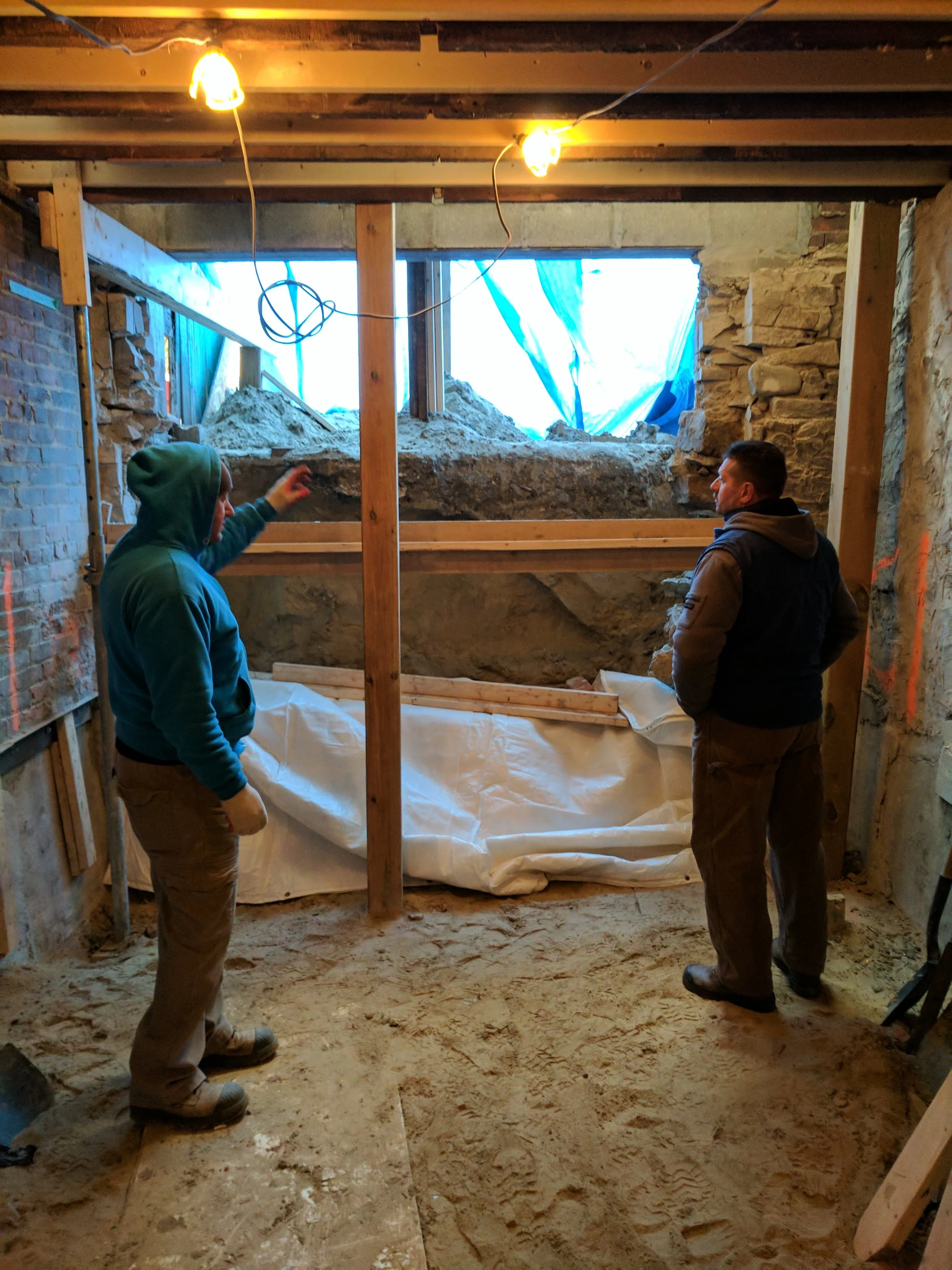 Two men working in a construction site, inspecting a partially excavated wall with sand on the ground and wooden support beams.