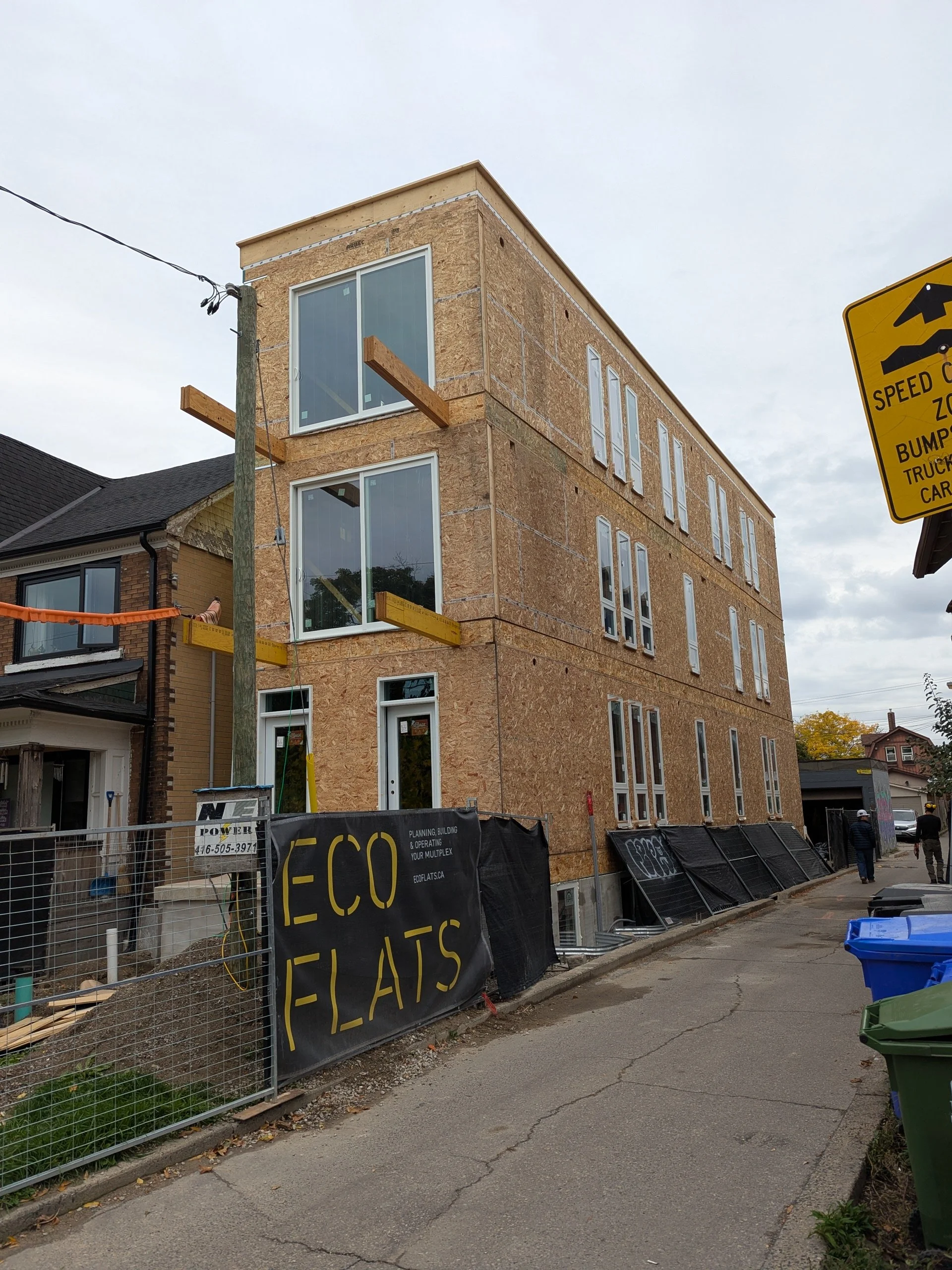 New multi-story building under construction with plywood sheathing and windows, surrounded by construction fencing with a sign that reads 'ECO FLATS'.