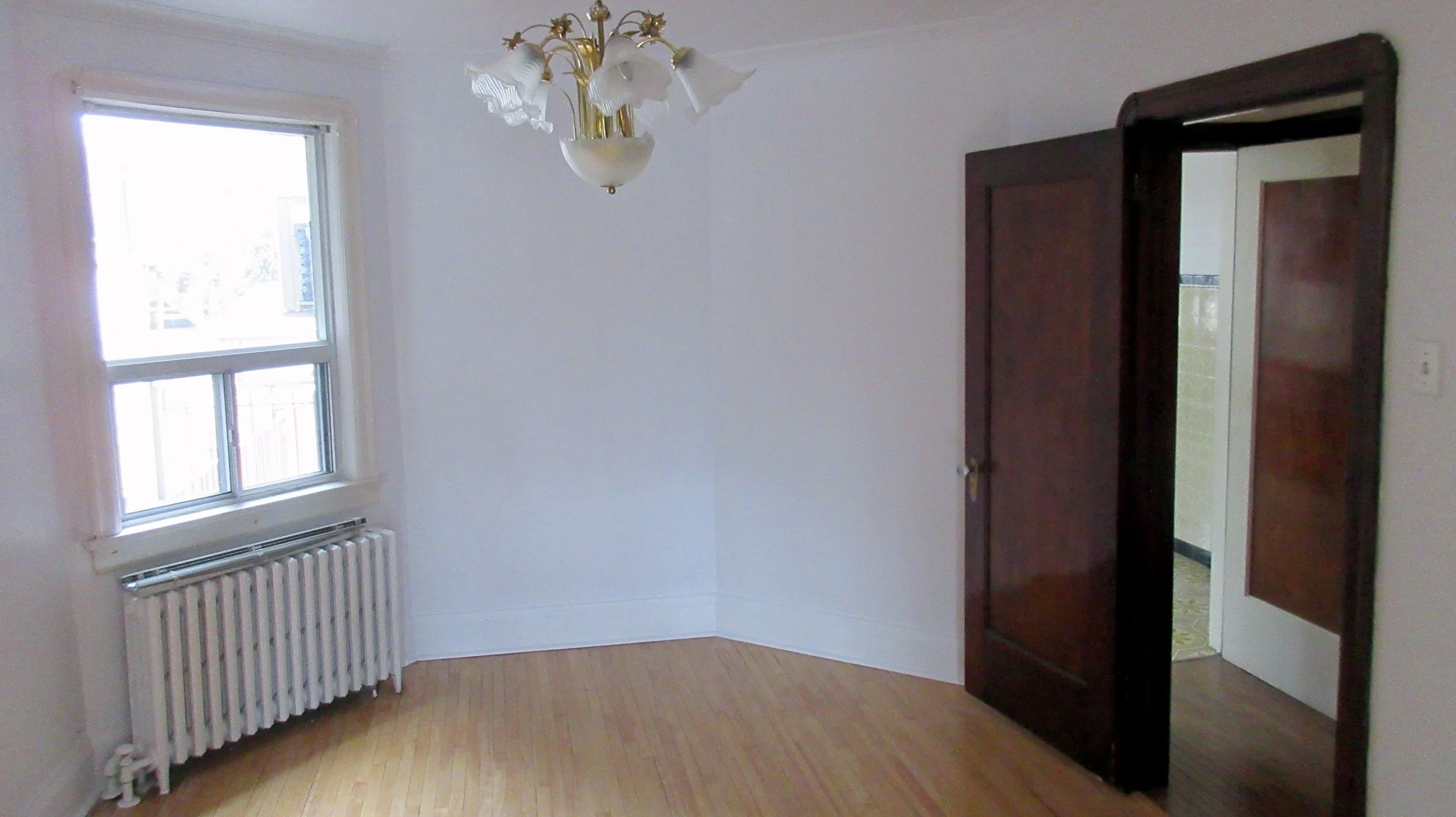 Empty room with wooden floor, a window with white trim, a radiator underneath, a hanging chandelier, and a dark wooden closet door partially open showing a room beyond.